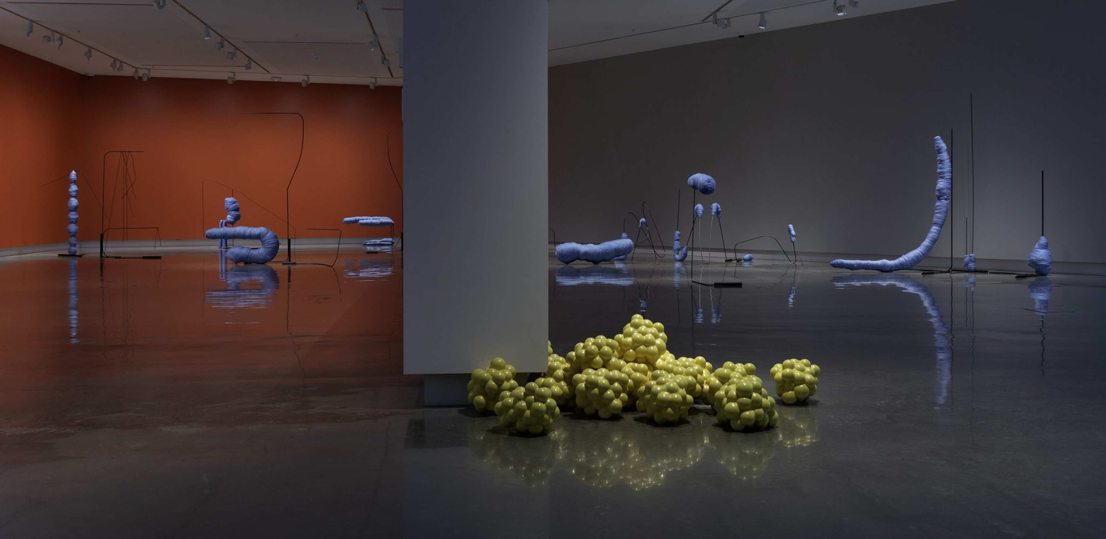A contemporary art gallery with scattered abstract sculptures: pale purple organic shapes on thin stands and a cluster of yellow, grape-like forms on the floor near a central pillar. The walls are orange and gray, and the floor is polished concrete.