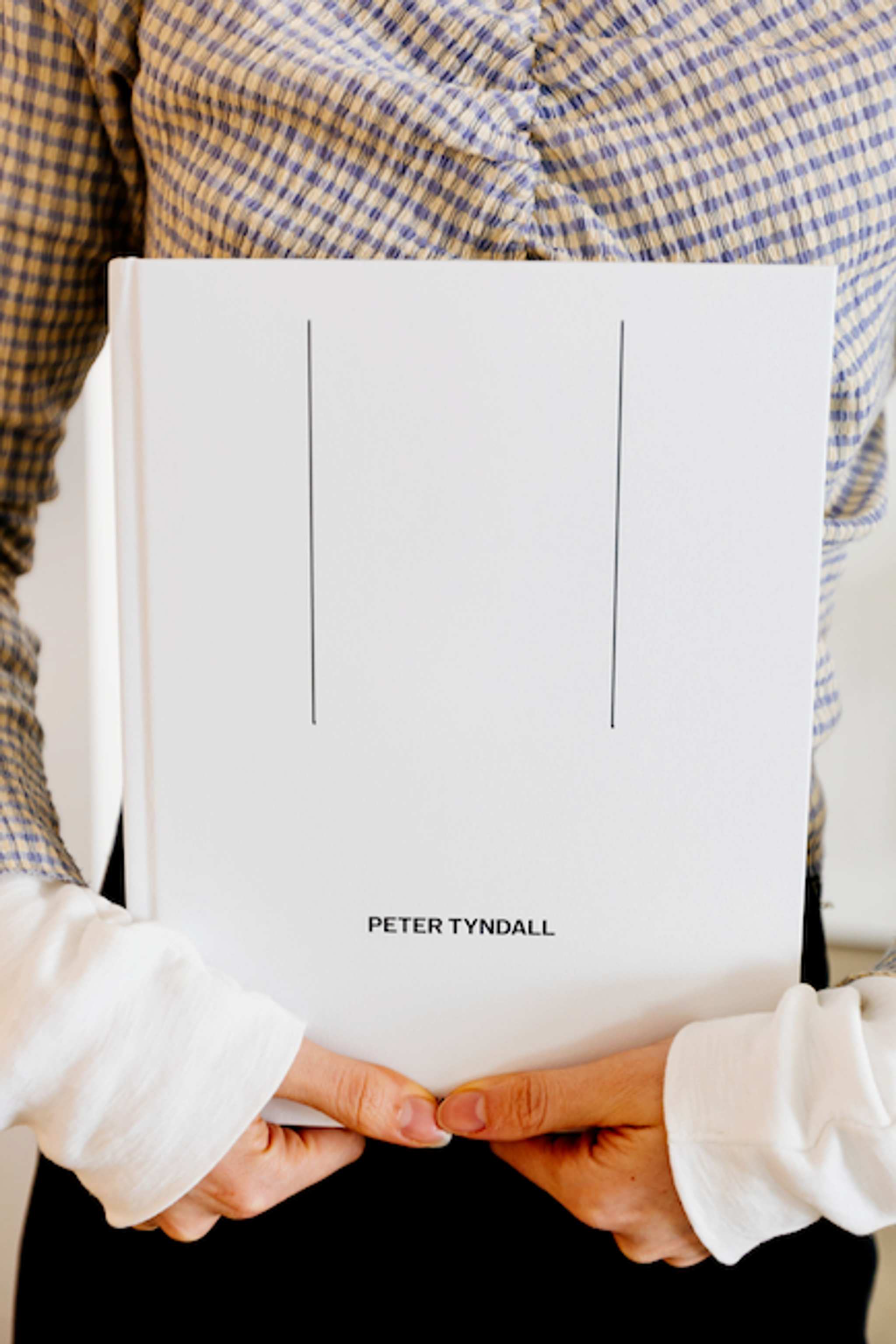 A person wearing a checkered shirt and white sleeves holds a white book titled PETER TYNDALL with two thin vertical lines on the cover.