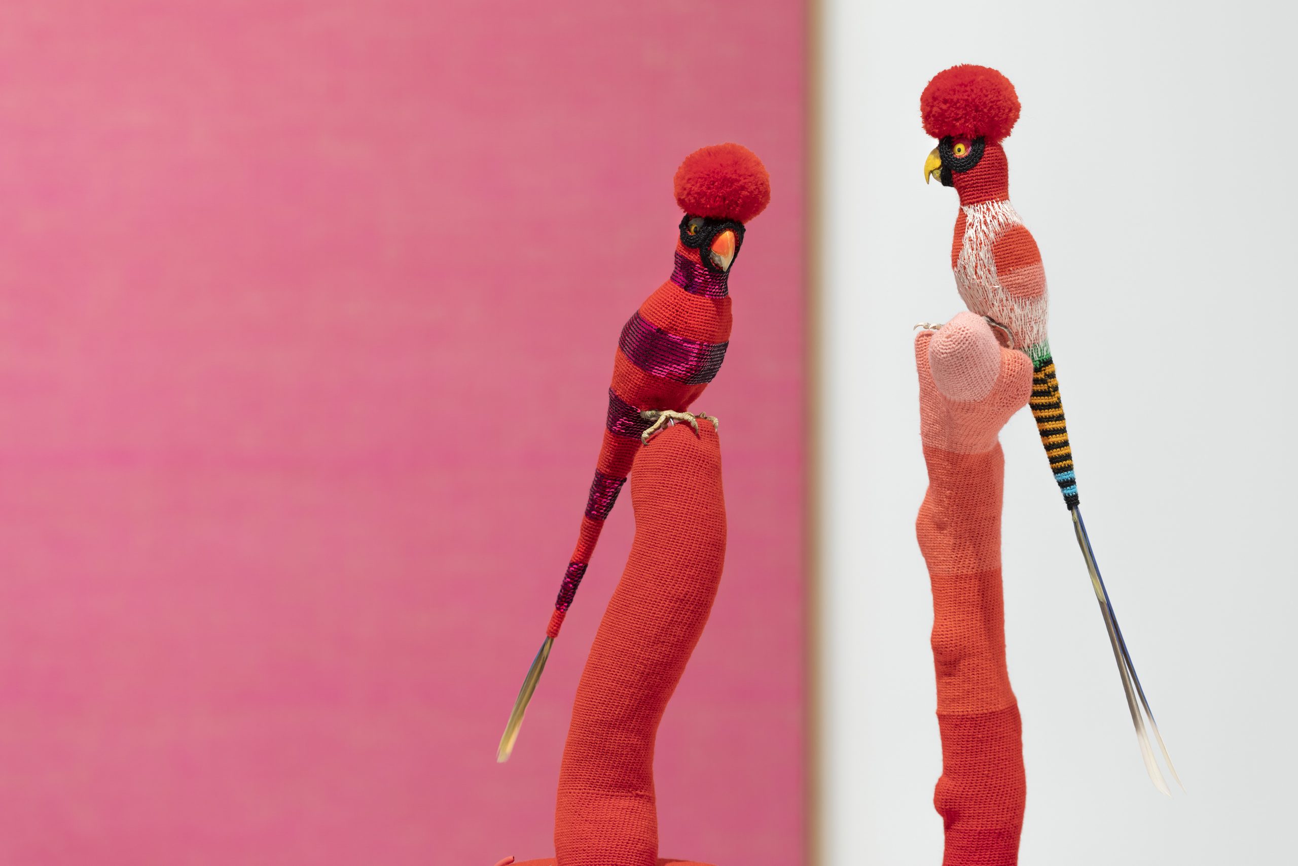 Two colorful bird sculptures with red pom-pom crests perch on red textile-covered stands, set against a blurred pink and white background.