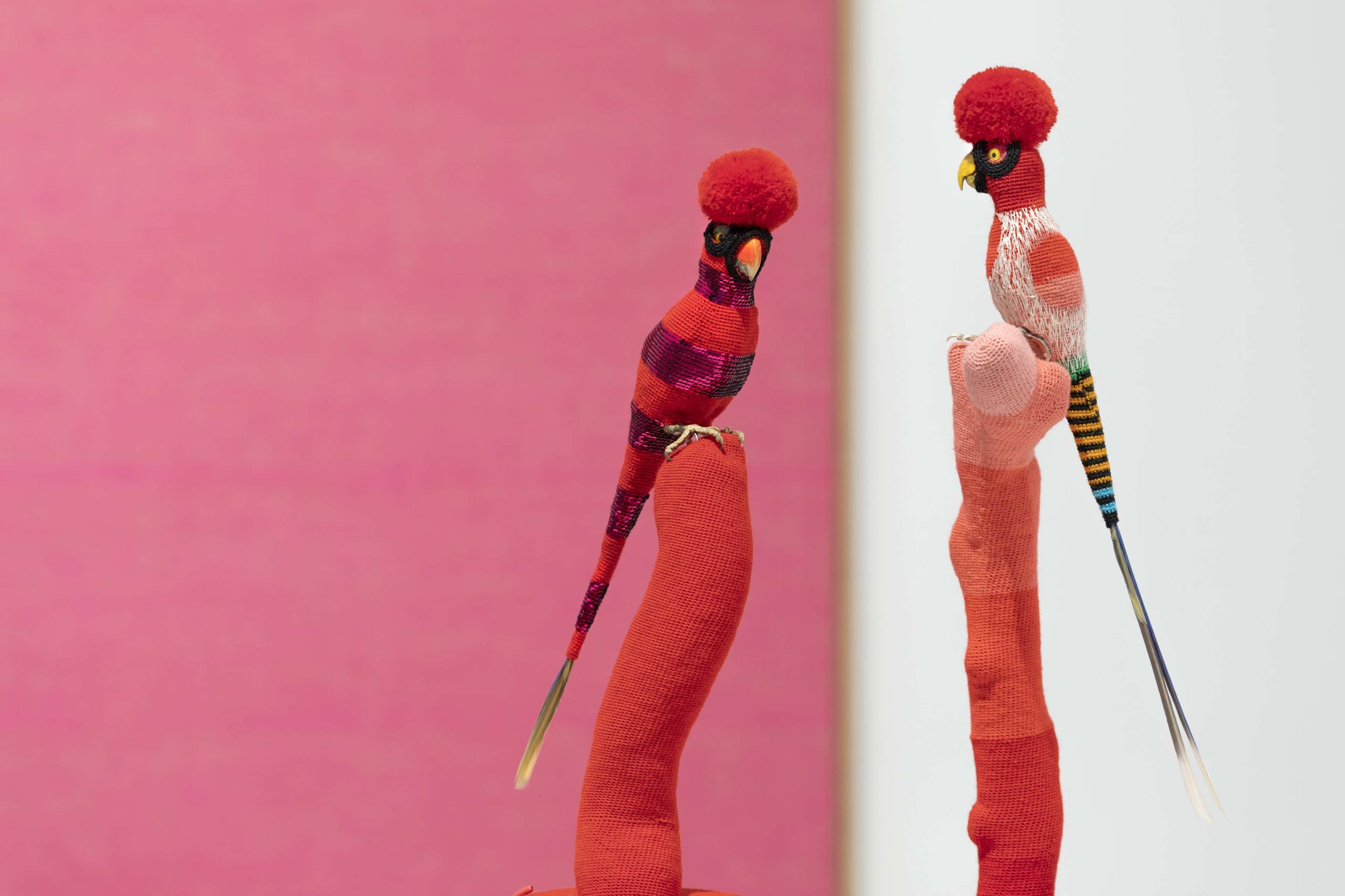 Two colorful bird sculptures with red pom-pom crests perch on red textile-covered stands, set against a blurred pink and white background.