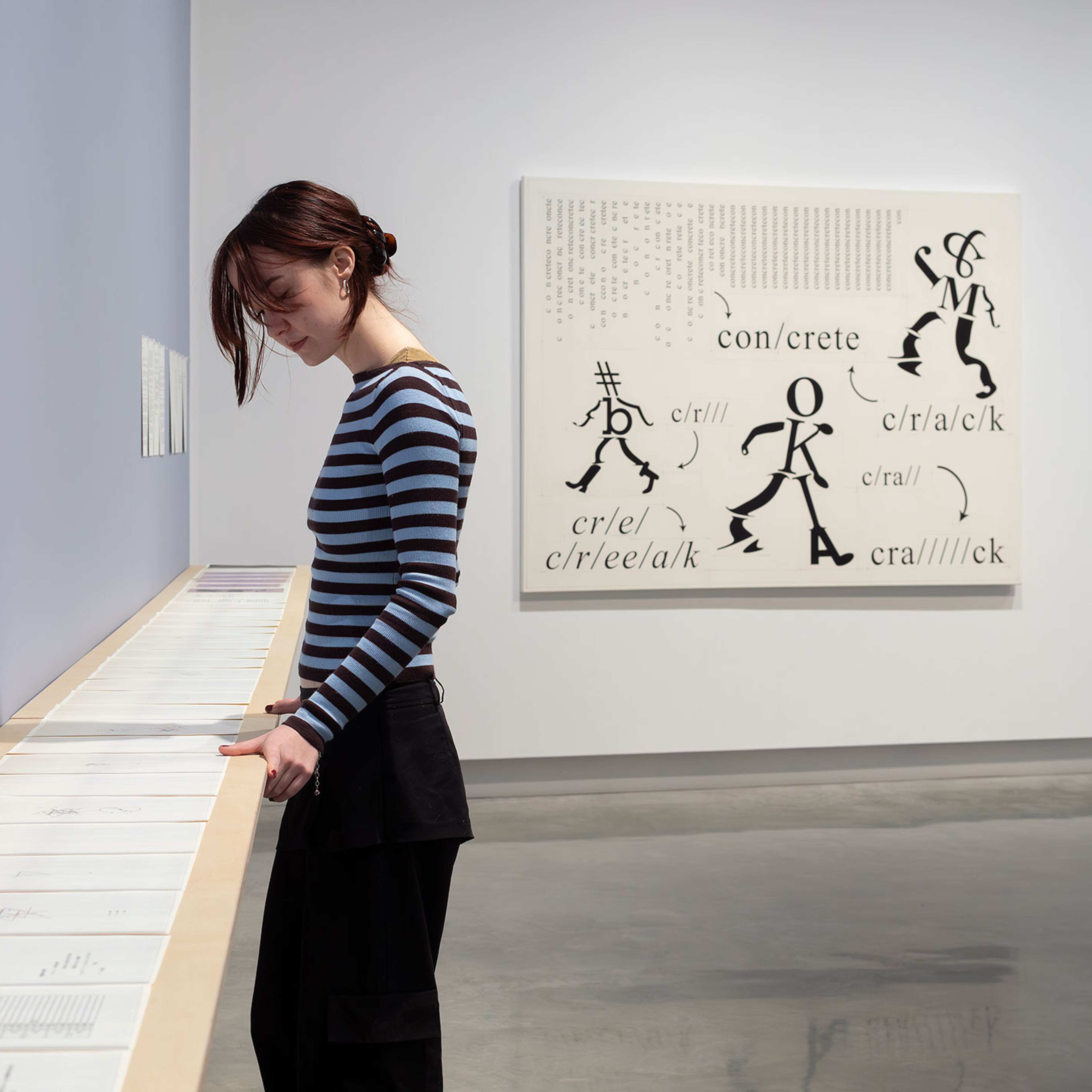 A person in a blue and black top looks down at a cabines of artworks in a contemporary gallery space with abstract black and white artwork in background