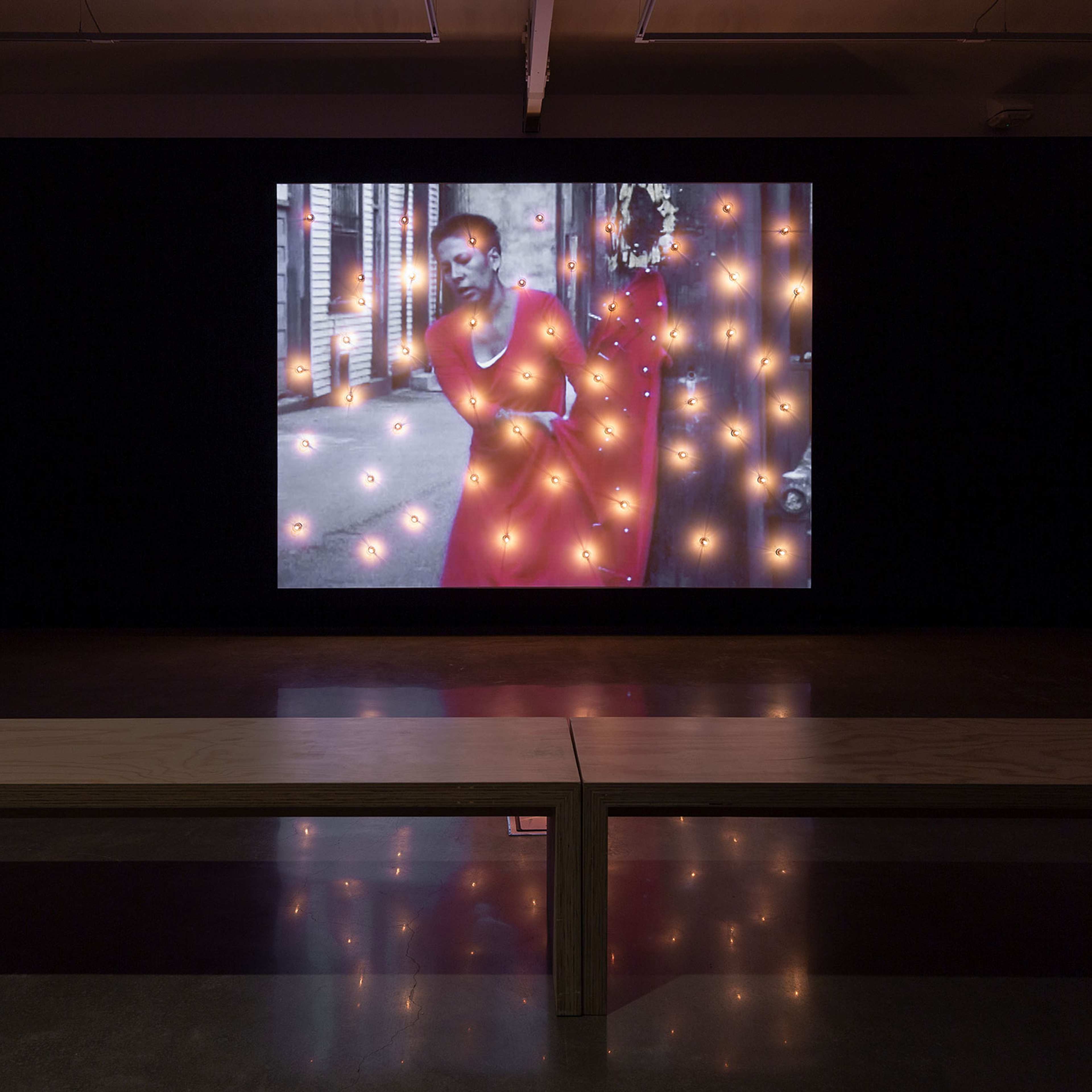 A wooden bench faces a large screen in a dark room. The screen displays a person in a red outfit with glowing orange dots scattered across the image, creating an artistic and cinematic effect.