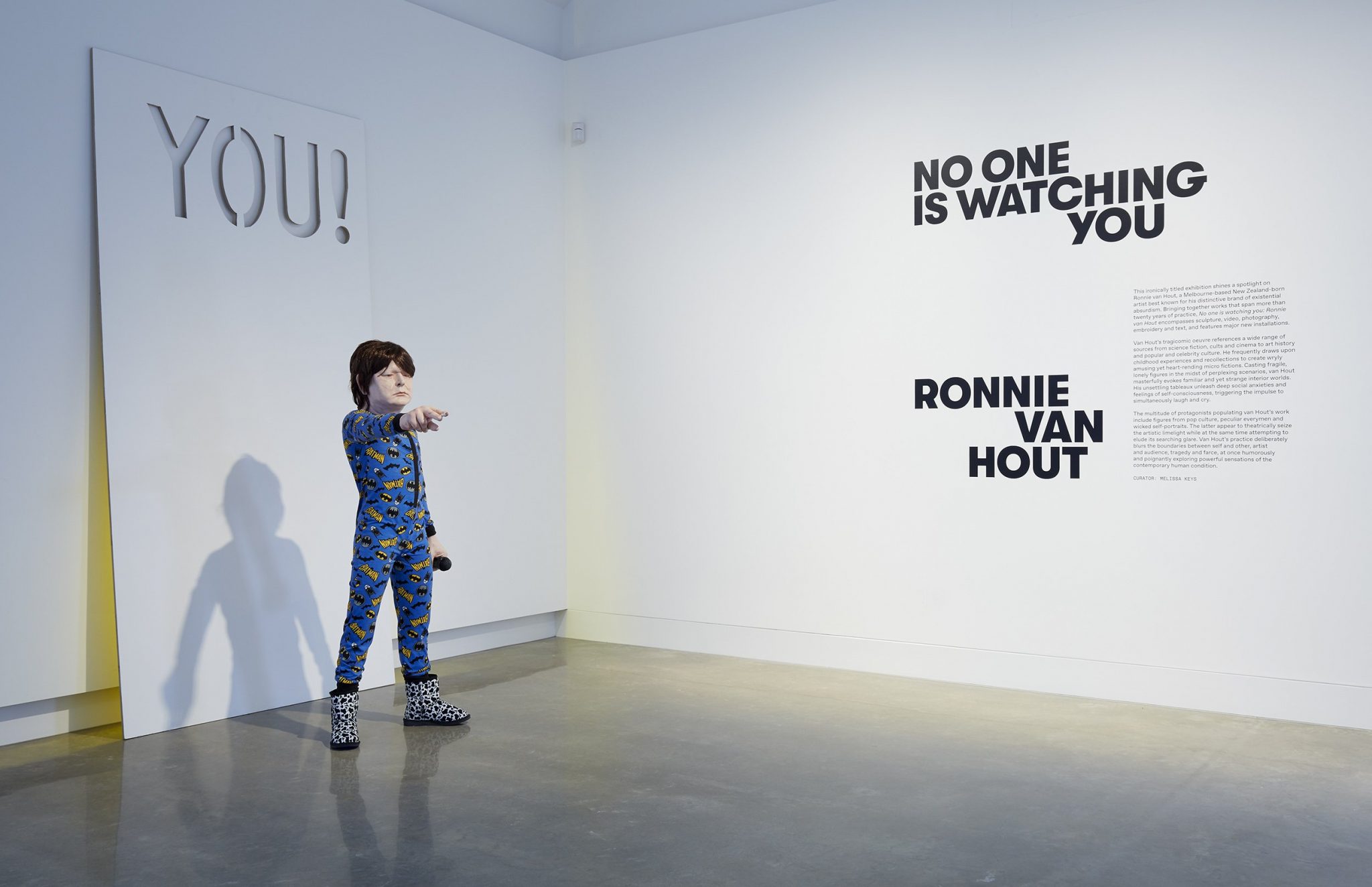 A child-sized mannequin in pajamas and animal print shoes stands in a gallery, pointing toward the camera. On the wall are bold black letters reading “NO ONE IS WATCHING YOU” and “RONNIE VAN HOUT.”.
