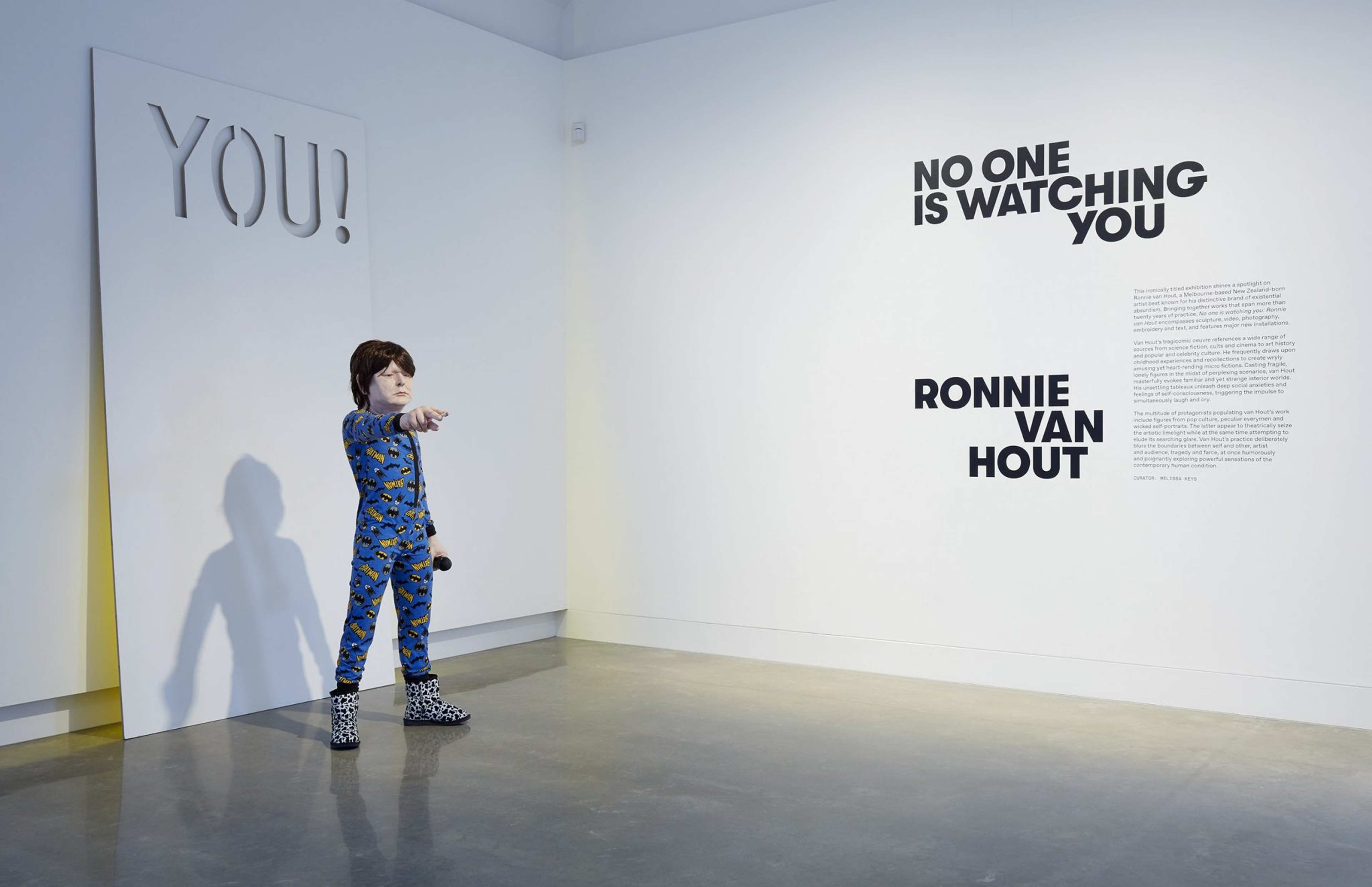 A child-sized mannequin in pajamas and animal print shoes stands in a gallery, pointing toward the camera. On the wall are bold black letters reading “NO ONE IS WATCHING YOU” and “RONNIE VAN HOUT.”.