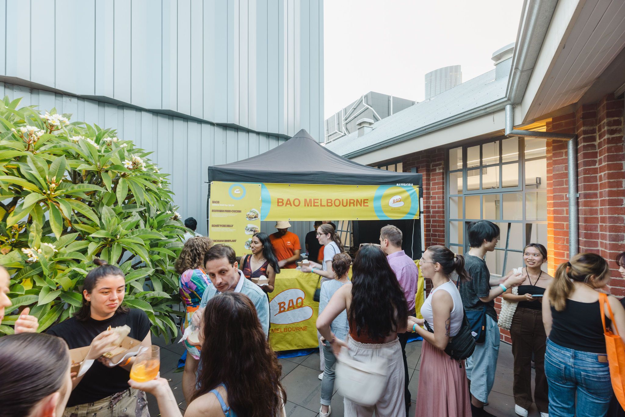 A group of people socialises and enjoys food and drinks in an outdoor courtyard near a yellow BAO MELBOURNE food stall under a canopy. Brick and metal buildings surround the area, and a green tree is visible on the left.