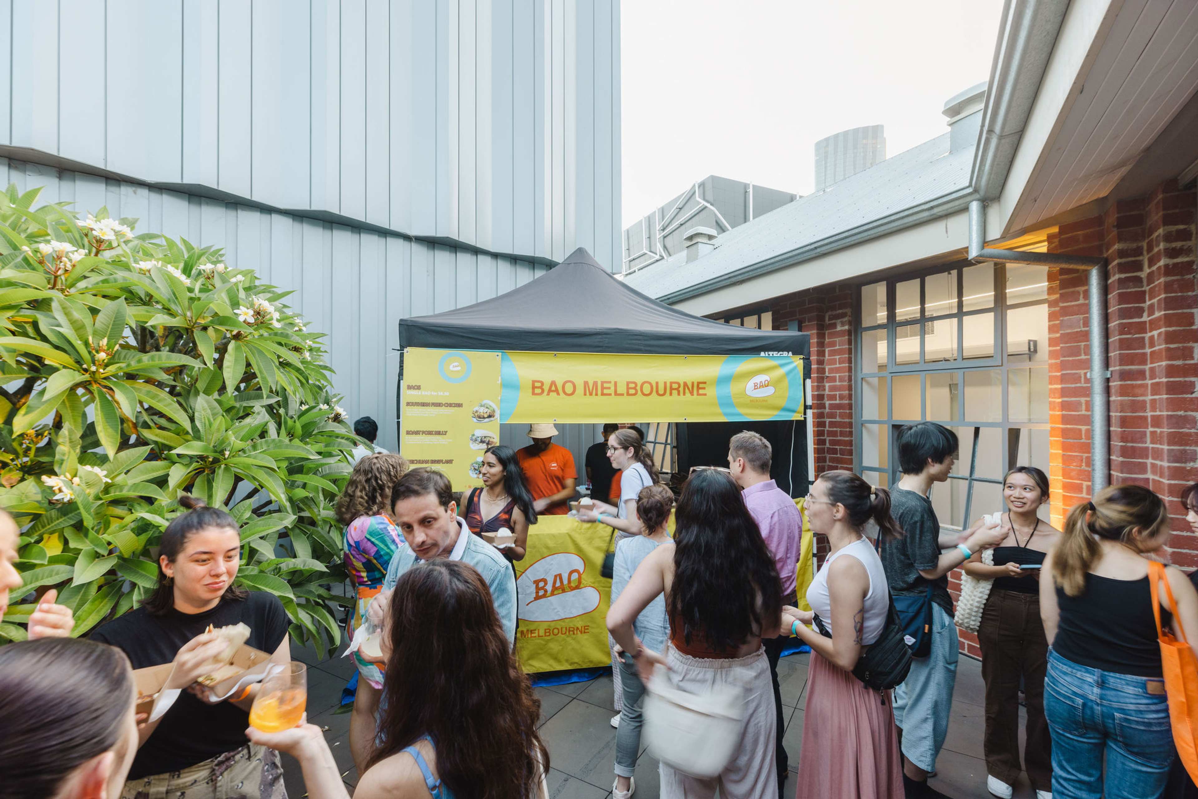 A group of people socialises and enjoys food and drinks in an outdoor courtyard near a yellow BAO MELBOURNE food stall under a canopy. Brick and metal buildings surround the area, and a green tree is visible on the left.