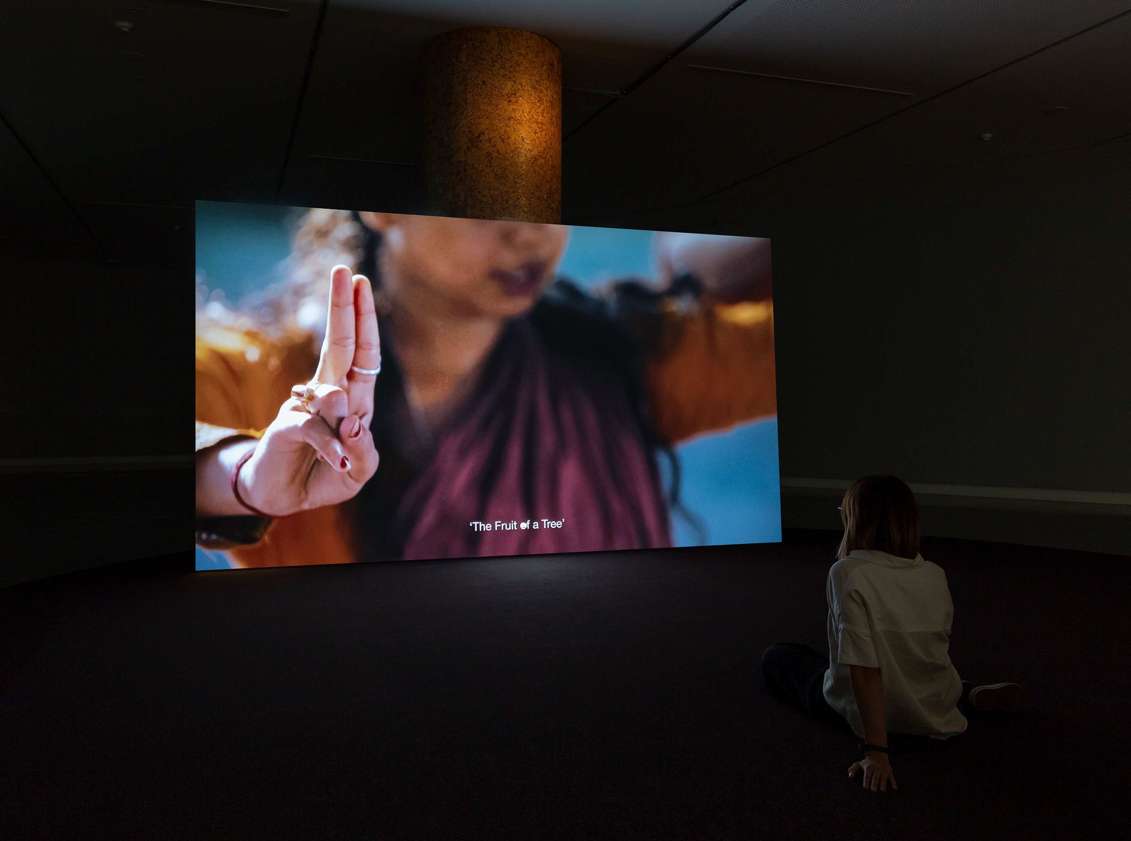A person sits on the floor in a dark room, watching a large screen displaying a woman making a hand gesture with two fingers raised. The screens subtitle reads, The Fruit will be Two.