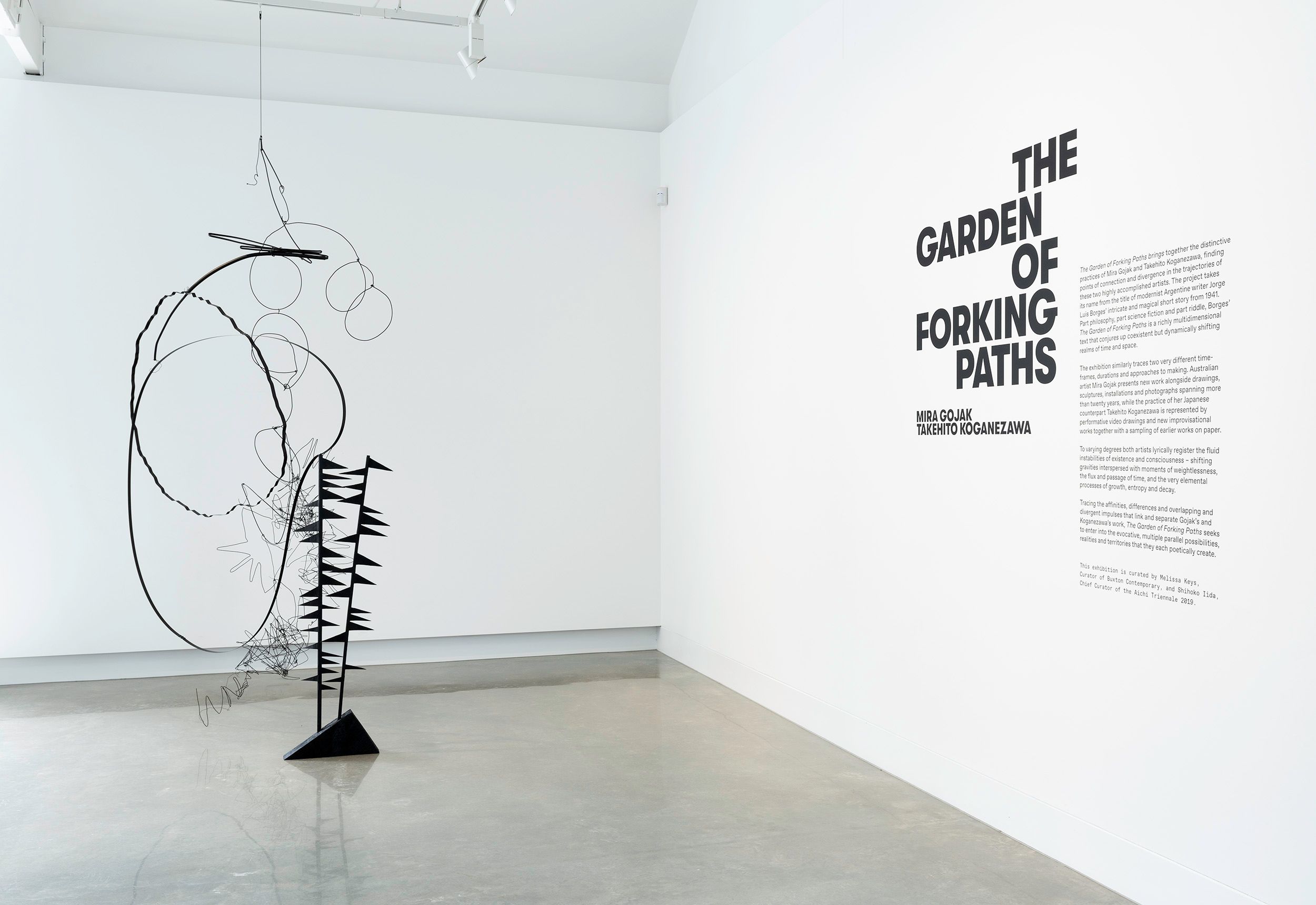 A minimalist gallery space features a large abstract black wire sculpture and a white wall with bold black text titled “The Garden of Forking Paths,” accompanied by an artist statement.