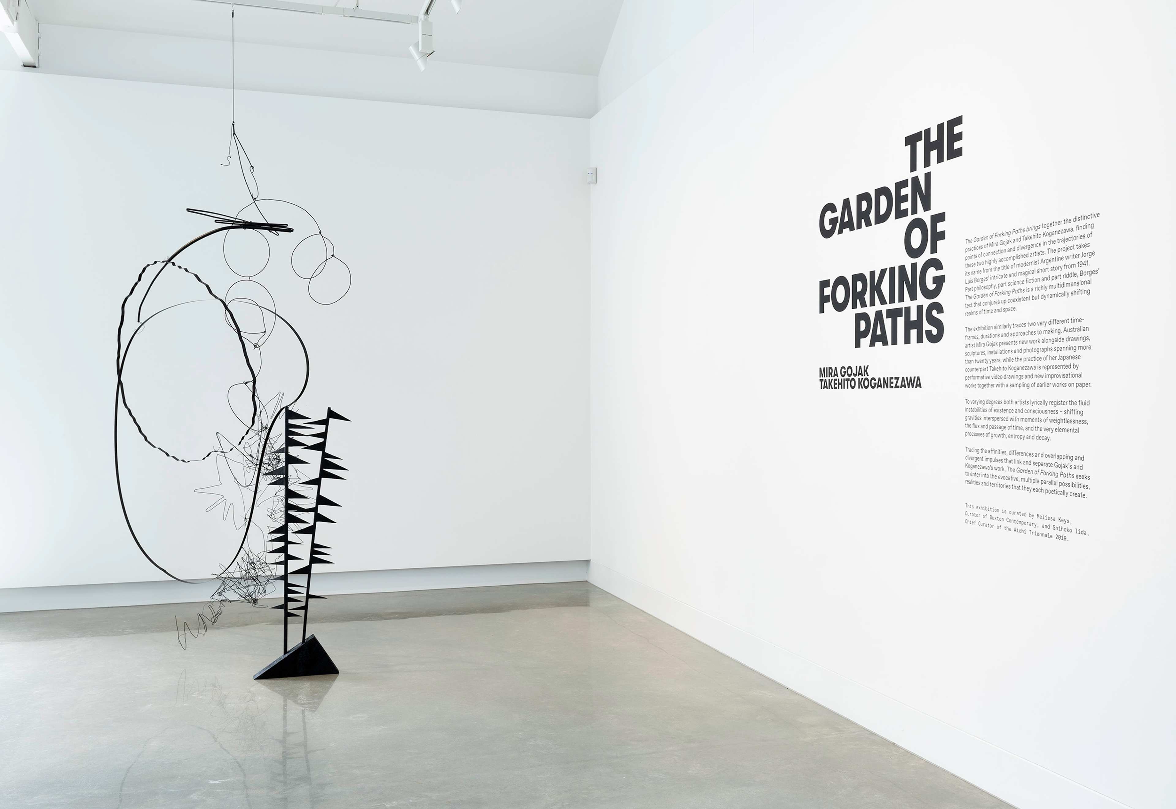 A minimalist gallery space features a large abstract black wire sculpture and a white wall with bold black text titled “The Garden of Forking Paths,” accompanied by an artist statement.