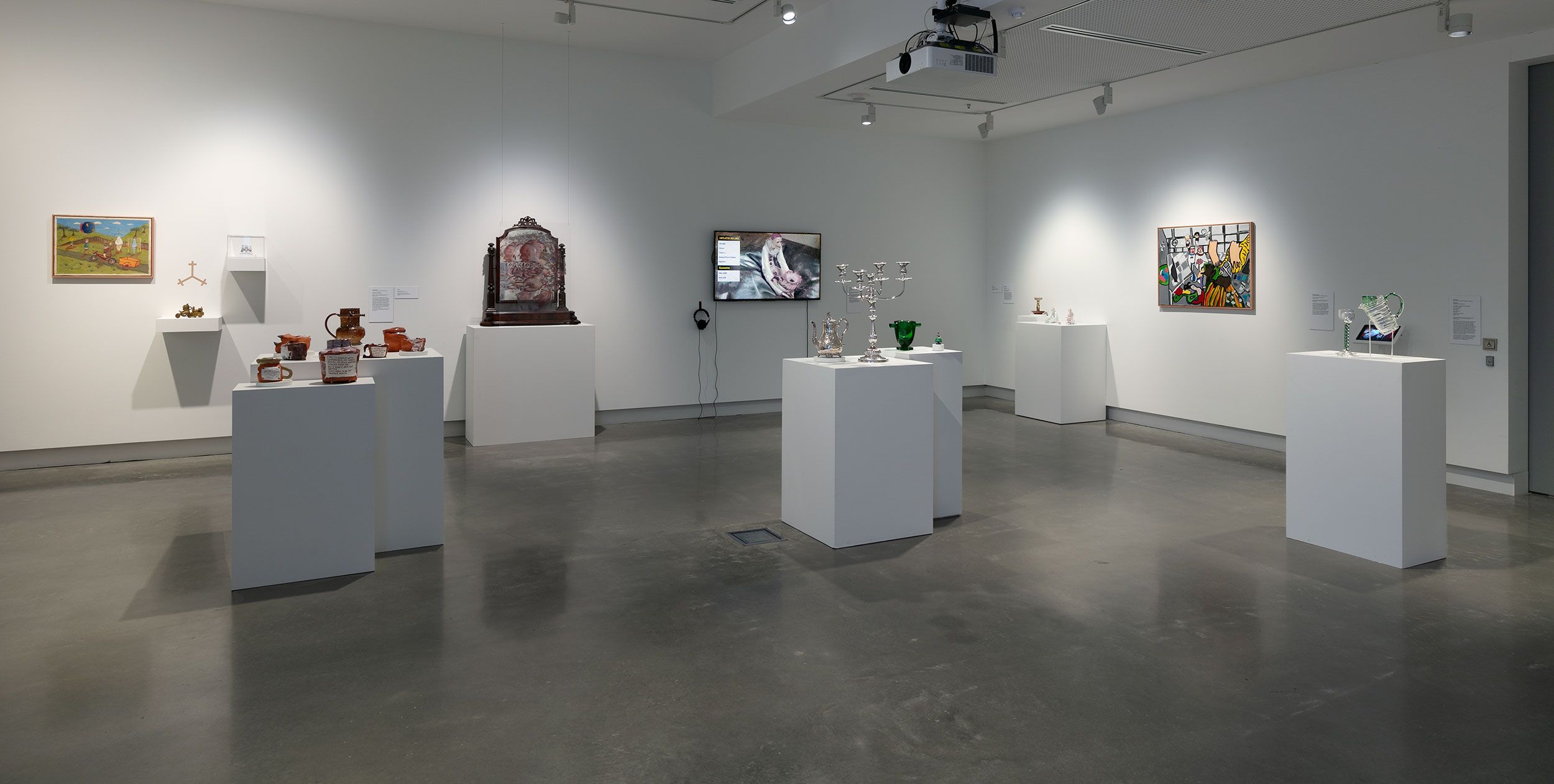 A modern art gallery room with white walls, featuring paintings and various sculptures displayed on white pedestals and mounted on the walls. The floor is gray concrete, and the space is brightly lit.