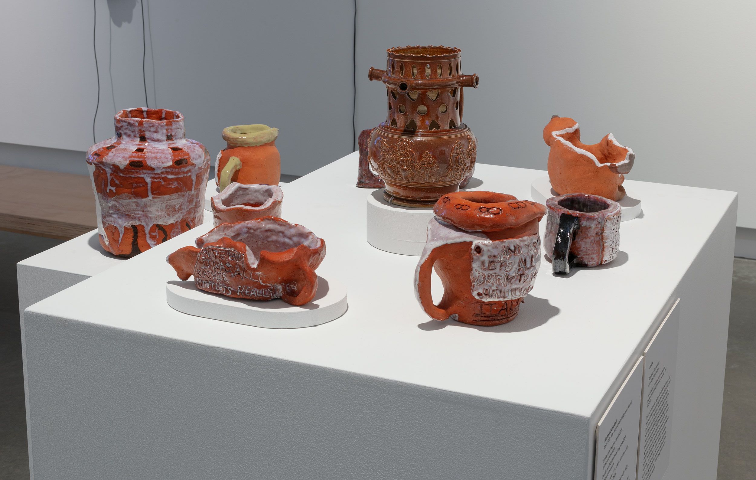 Seven ceramic vessels and sculptures are displayed on a white pedestal. The space is brightly lit and minimalist.