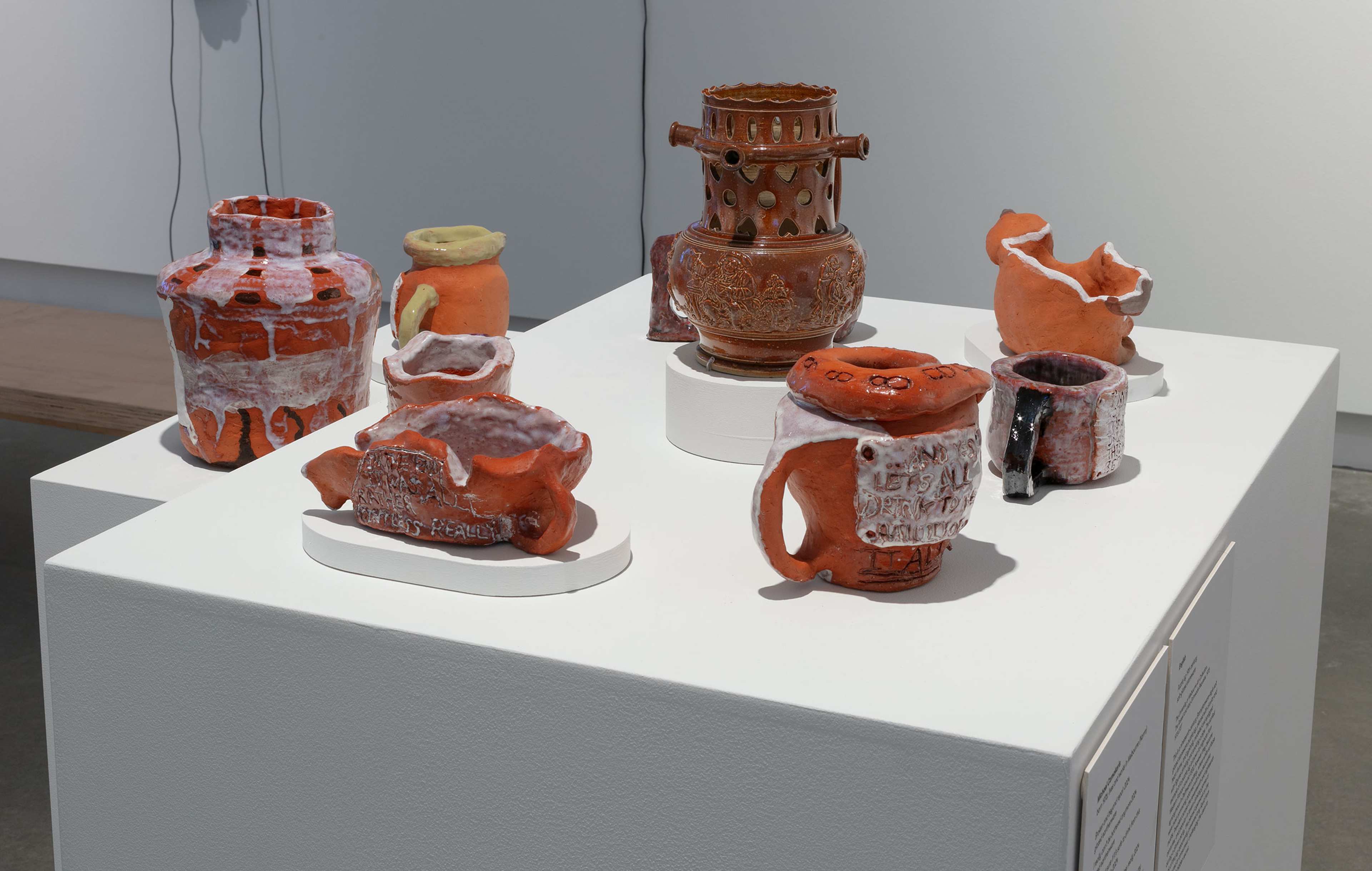 Seven ceramic vessels and sculptures are displayed on a white pedestal. The space is brightly lit and minimalist.