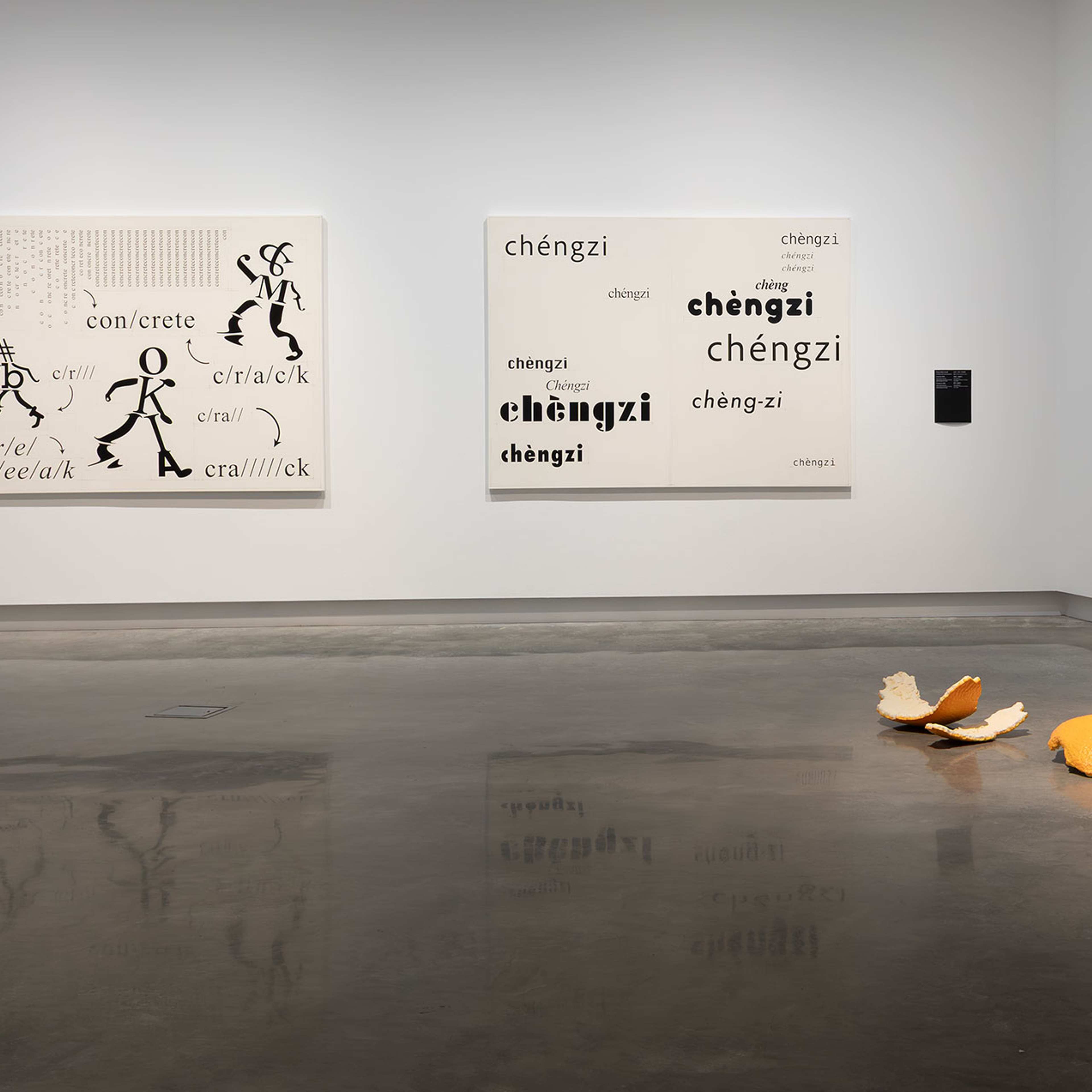 A contemporary art gallery with two text-based artworks on the wall and a large, sculptural orange peel displayed on the polished concrete floor. The space is clean and minimally decorated.