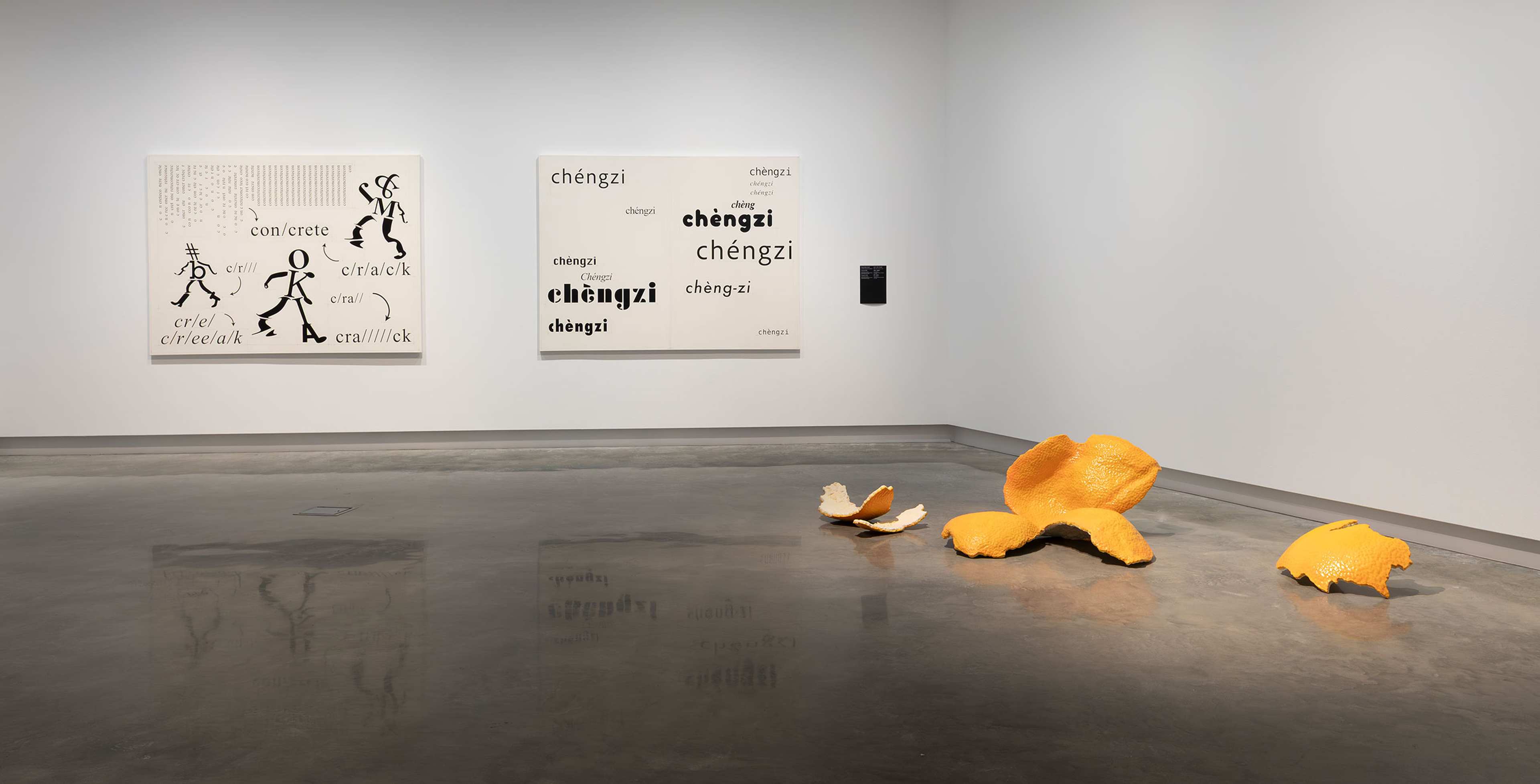 A contemporary art gallery with two text-based artworks on the wall and a large, sculptural orange peel displayed on the polished concrete floor. The space is clean and minimally decorated.