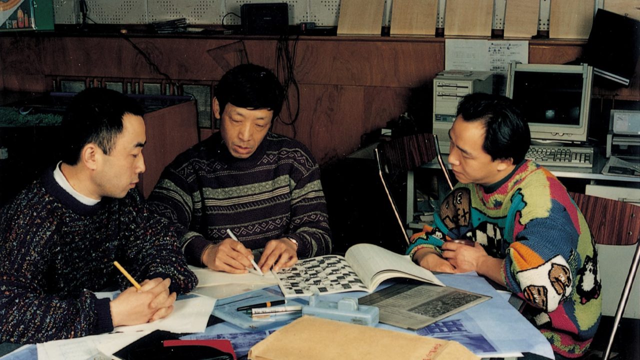 Three men sit at a table covered with papers, books, and stationery, engaged in discussion or study. They appear focused, with one man pointing at a printed page. Computers and shelves are seen in the background.