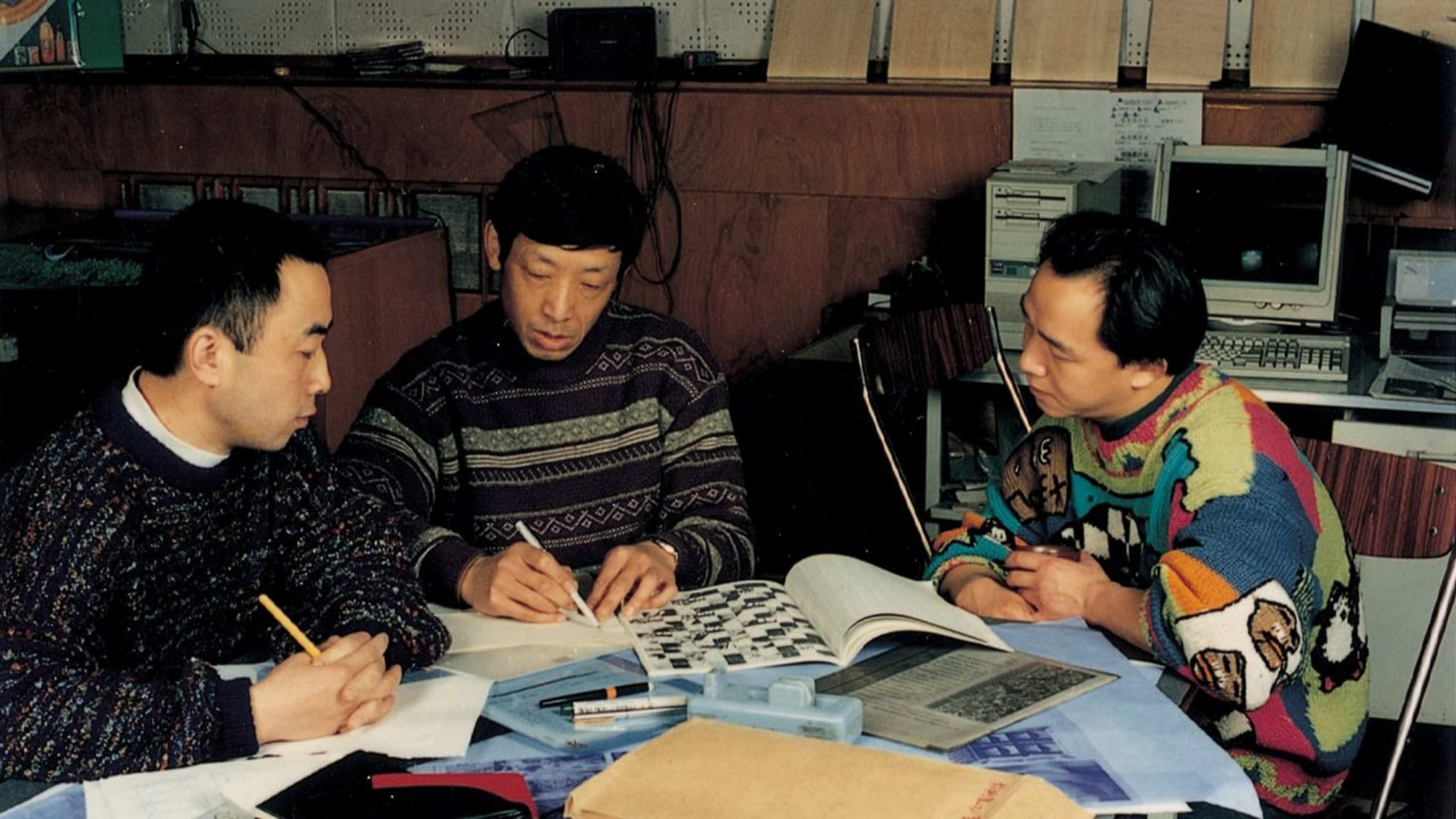 Three men sit at a table covered with papers, books, and stationery, engaged in discussion or study. They appear focused, with one man pointing at a printed page. Computers and shelves are seen in the background.