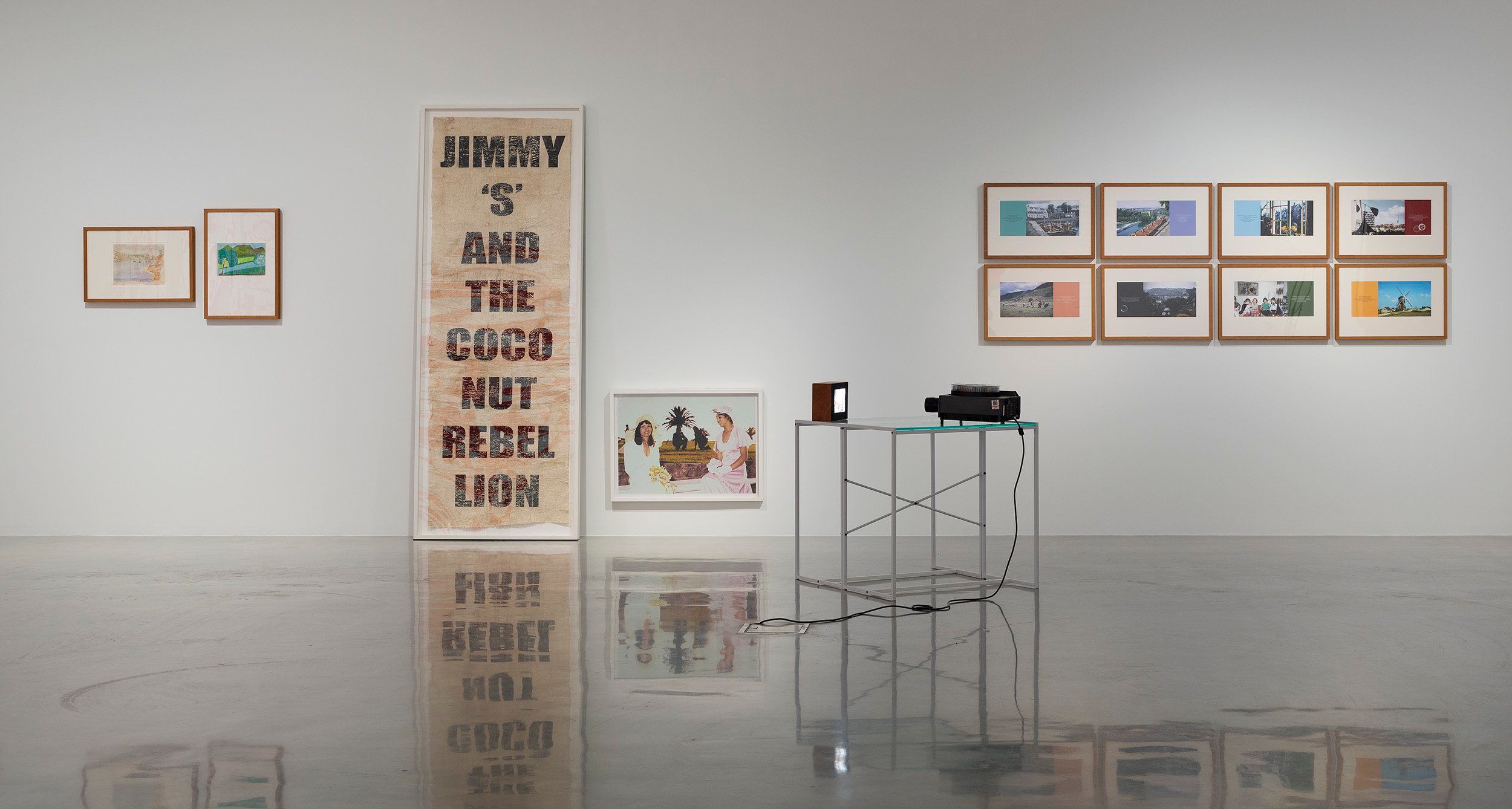 A contemporary art gallery with framed artworks on white walls, a large vertical piece reading JIMMY S AND THE COCONUT REBEL LION, and a glass table holding a projector with cables on a polished floor.