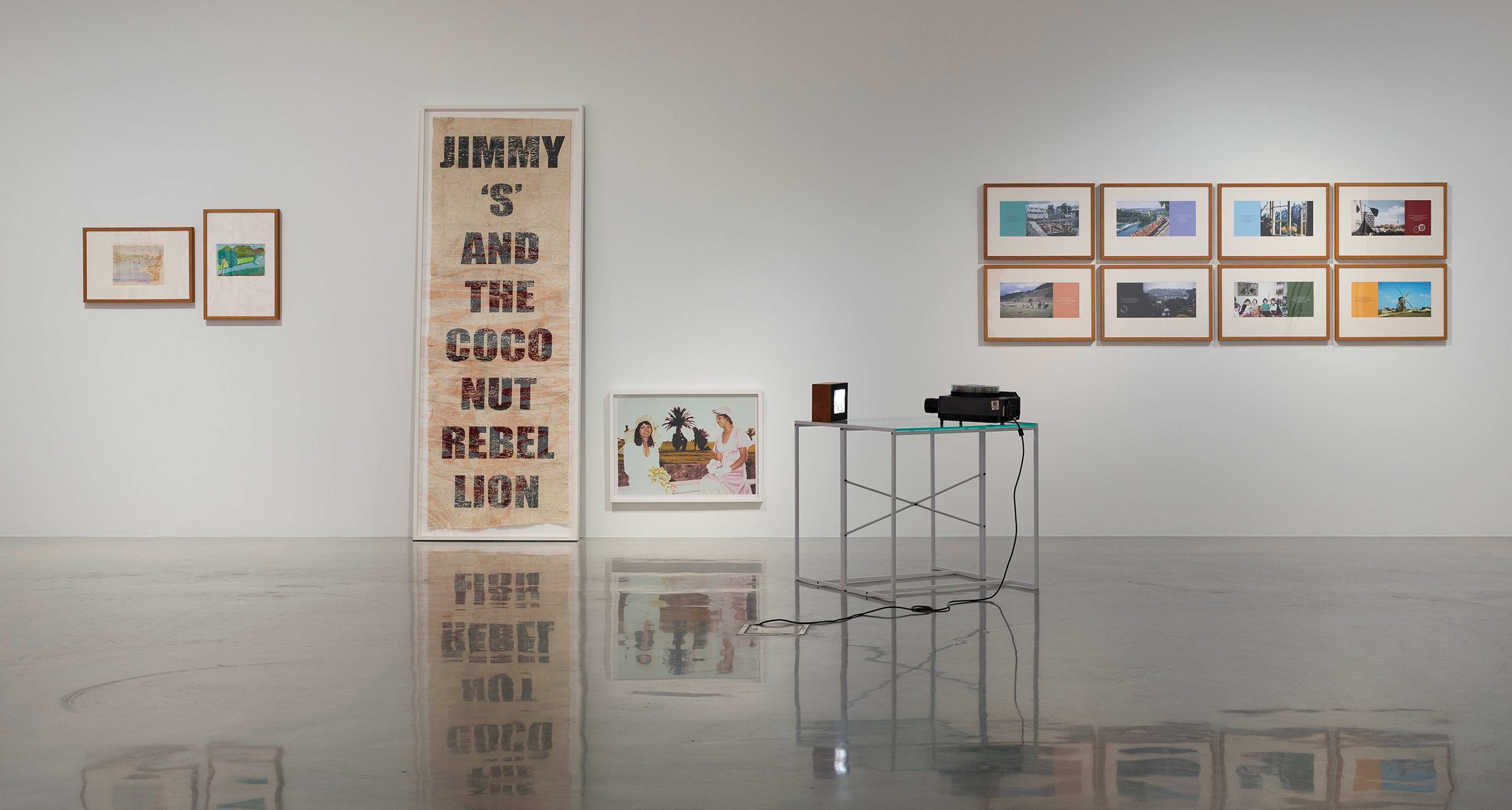 A contemporary art gallery with framed artworks on white walls, a large vertical piece reading JIMMY S AND THE COCONUT REBEL LION, and a glass table holding a projector with cables on a polished floor.