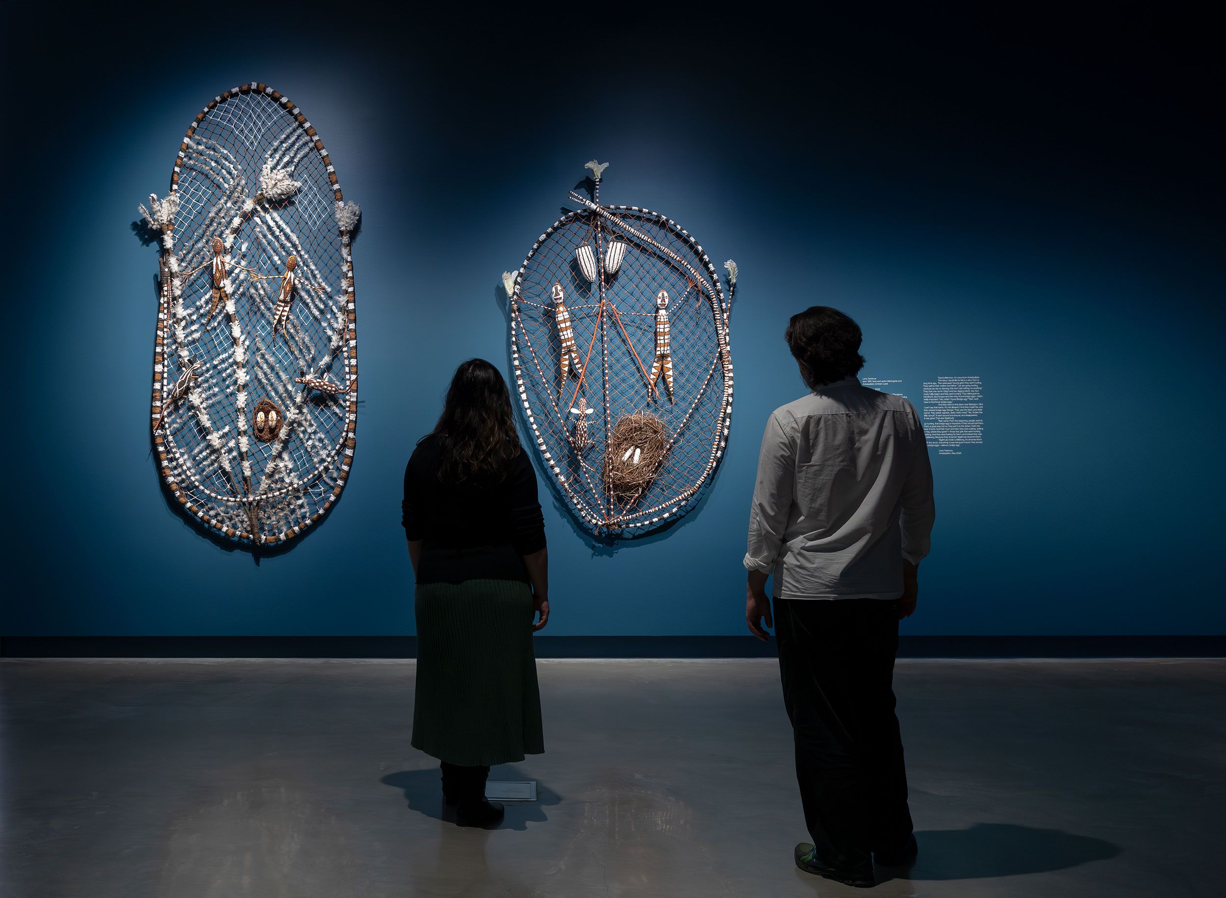 Two people stand in a dimly lit gallery, facing two large, intricate woven artworks displayed on a deep blue wall. The pieces feature shells, fibers, and natural materials arranged in oval shapes.