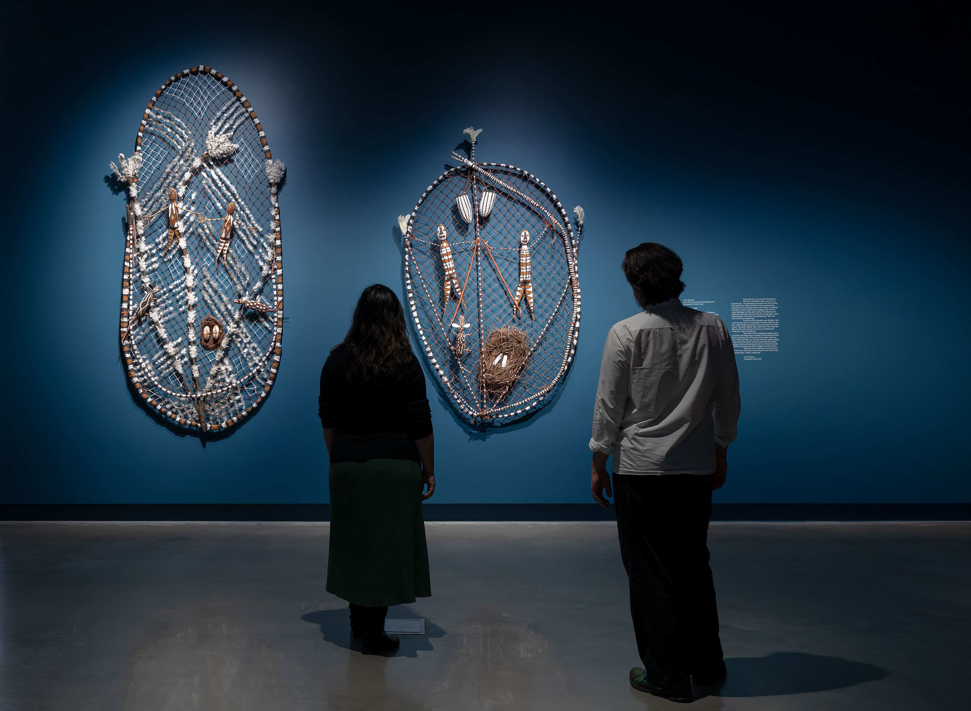 Two people stand in a dimly lit gallery, facing two large, intricate woven artworks displayed on a deep blue wall. The pieces feature shells, fibers, and natural materials arranged in oval shapes.
