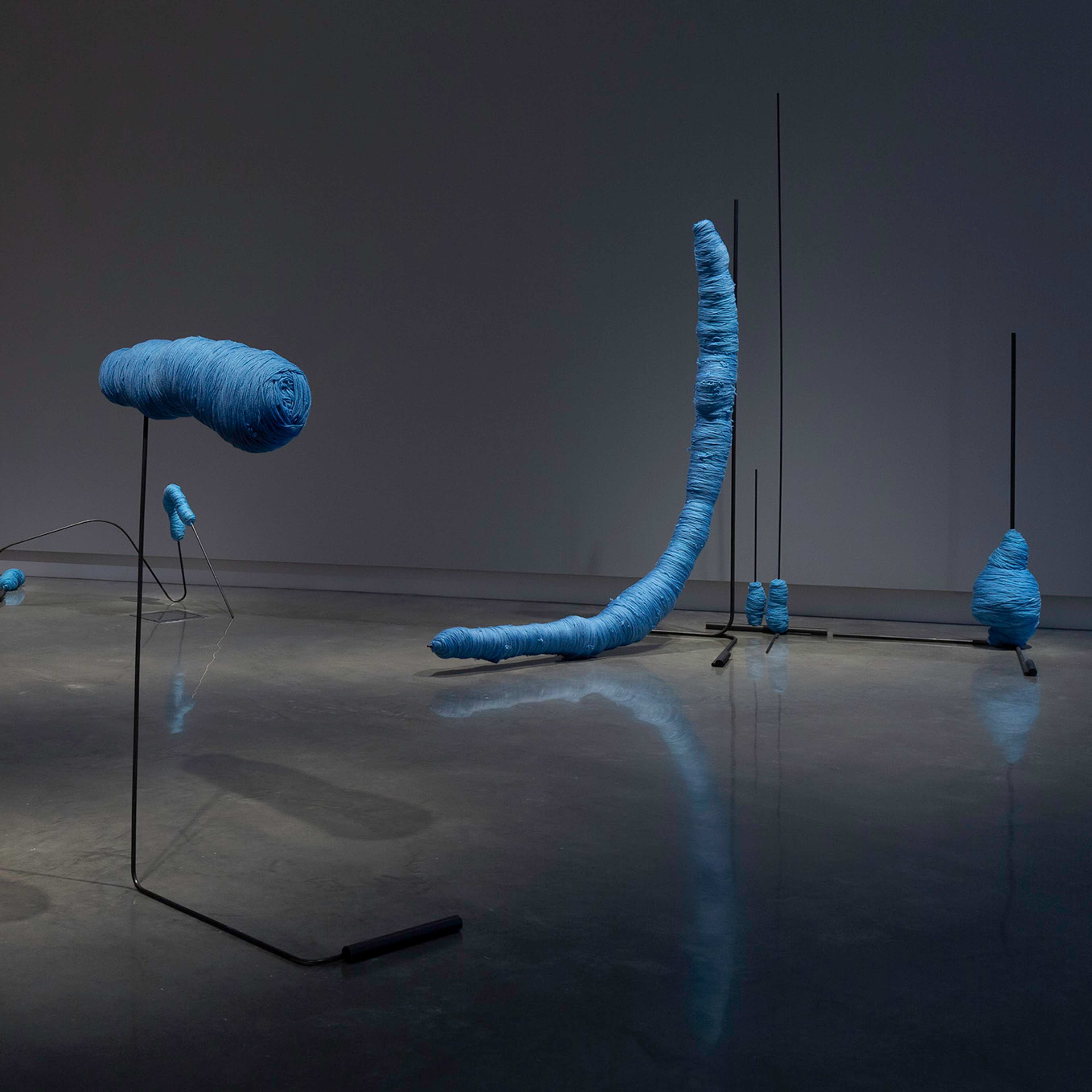 Contemporary art installation featuring abstract, elongated, blue forms wrapped in material, mounted on thin, black metal stands of varying heights, arranged across a shiny, reflective gallery floor under dim lighting.