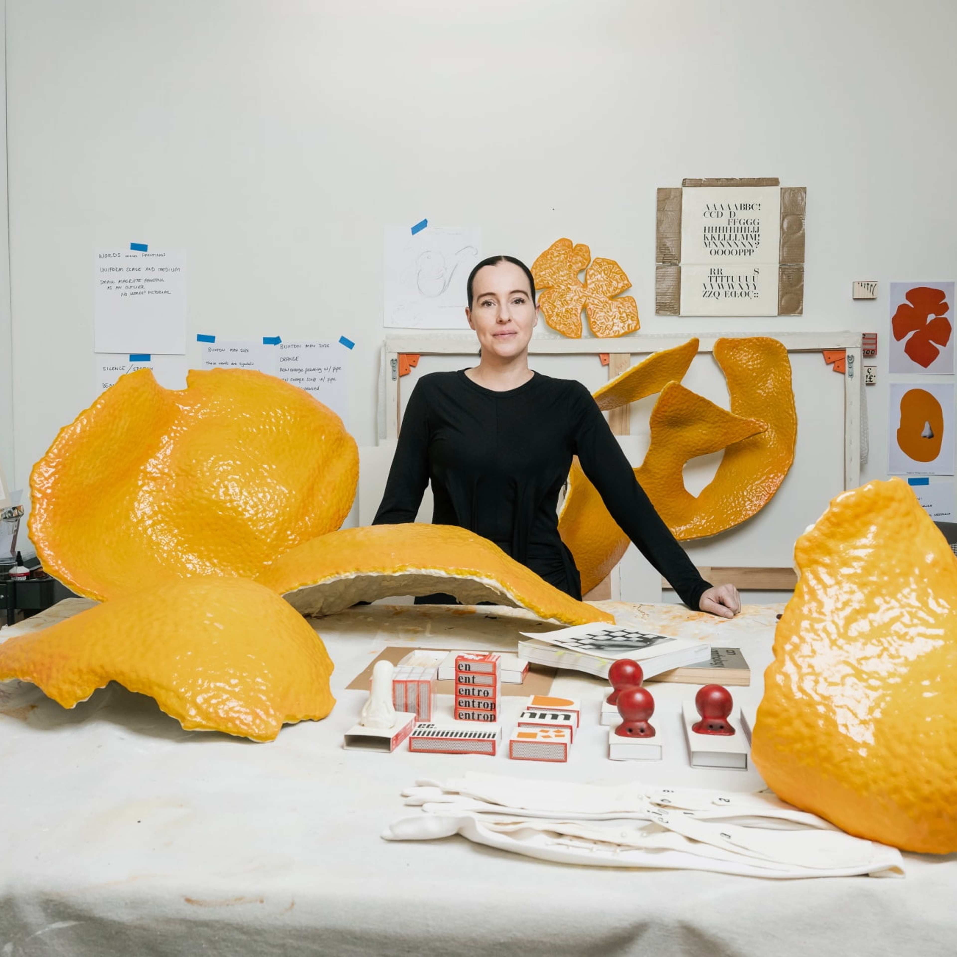 A person stands in an art studio behind a table covered with orange sculptural pieces, stamps, and art supplies. Artworks and images are displayed on the white walls in the background.