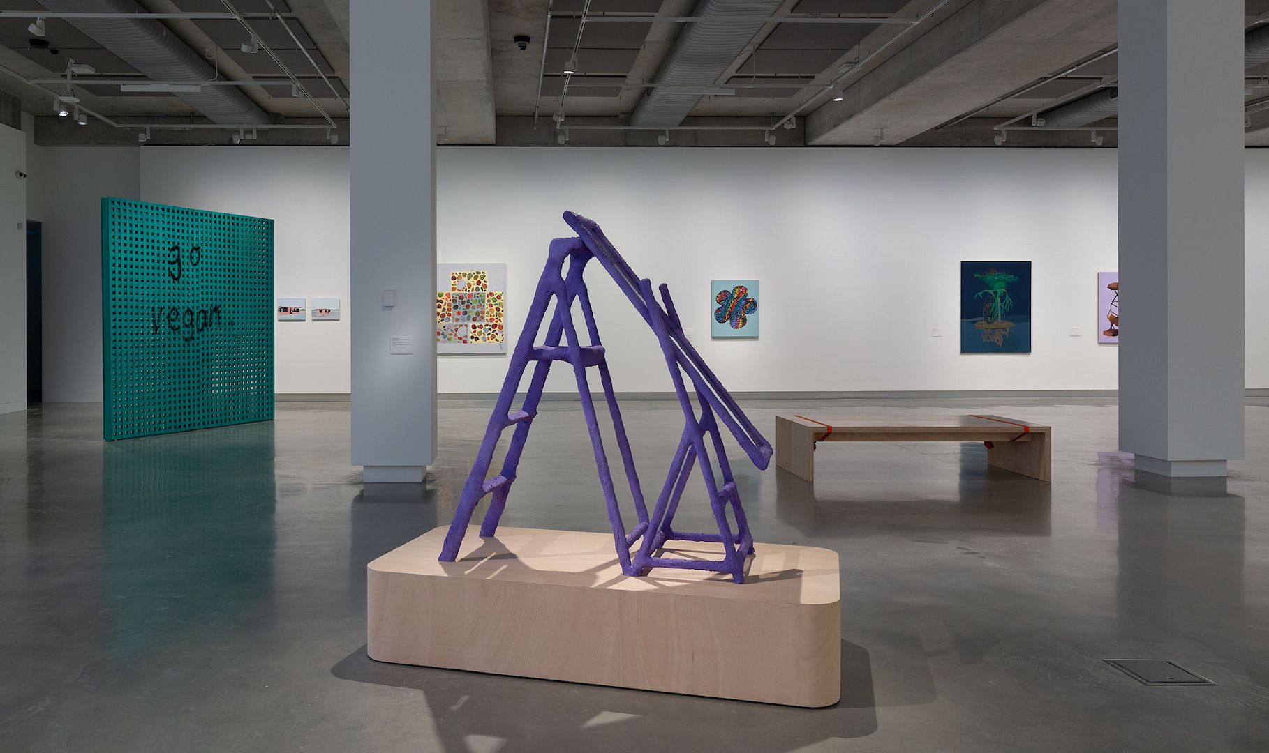 A contemporary art gallery with modern sculptures and paintings, including a central purple abstract structure on a wooden base, colorful wall art, and a simple wooden bench on a polished concrete floor.
