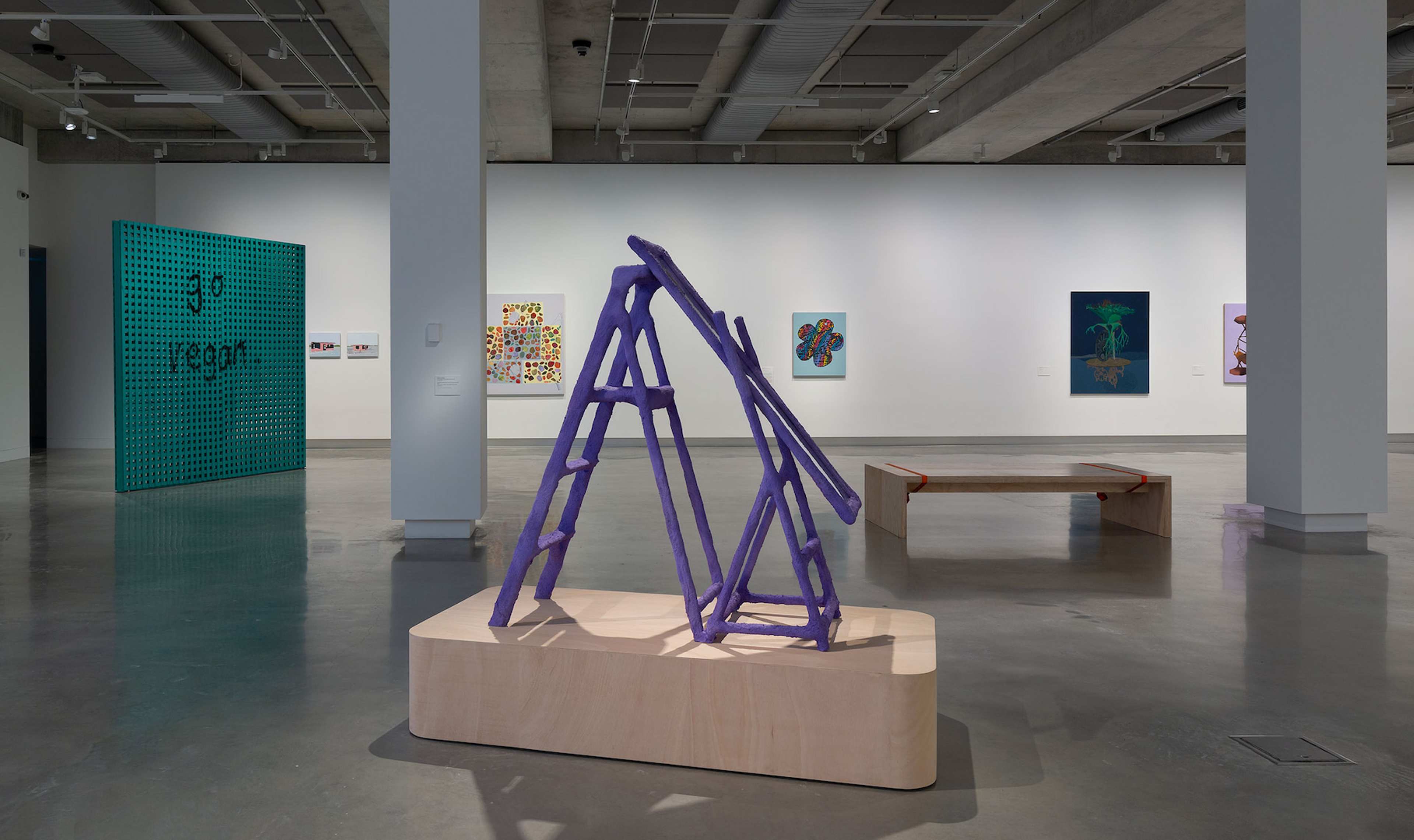 A contemporary art gallery with modern sculptures and paintings, including a central purple abstract structure on a wooden base, colorful wall art, and a simple wooden bench on a polished concrete floor.