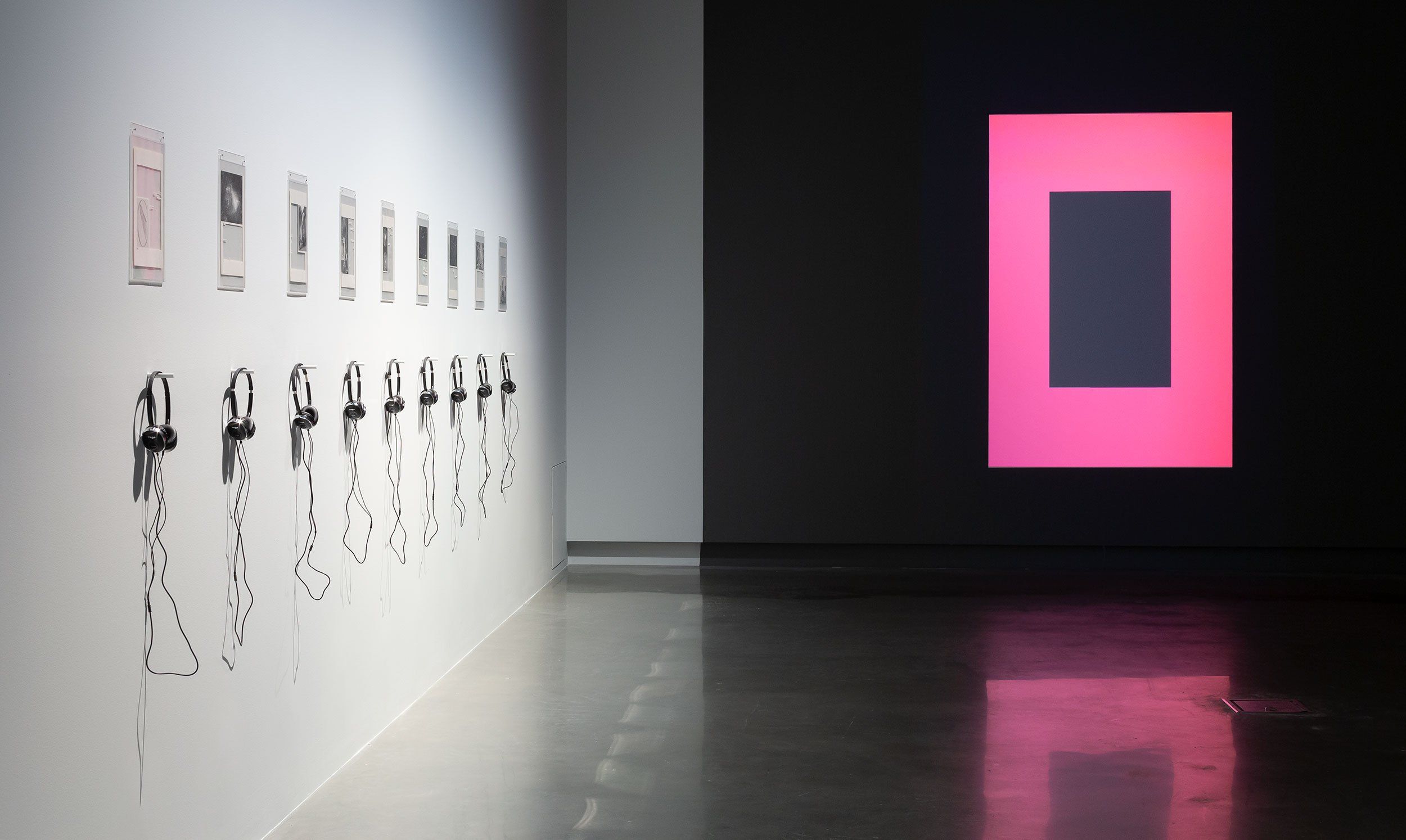 A modern gallery displays headphones hanging below framed images on a white wall, with a large glowing pink and black digital art piece projected on a dark wall in the background. The floor is reflective concrete.