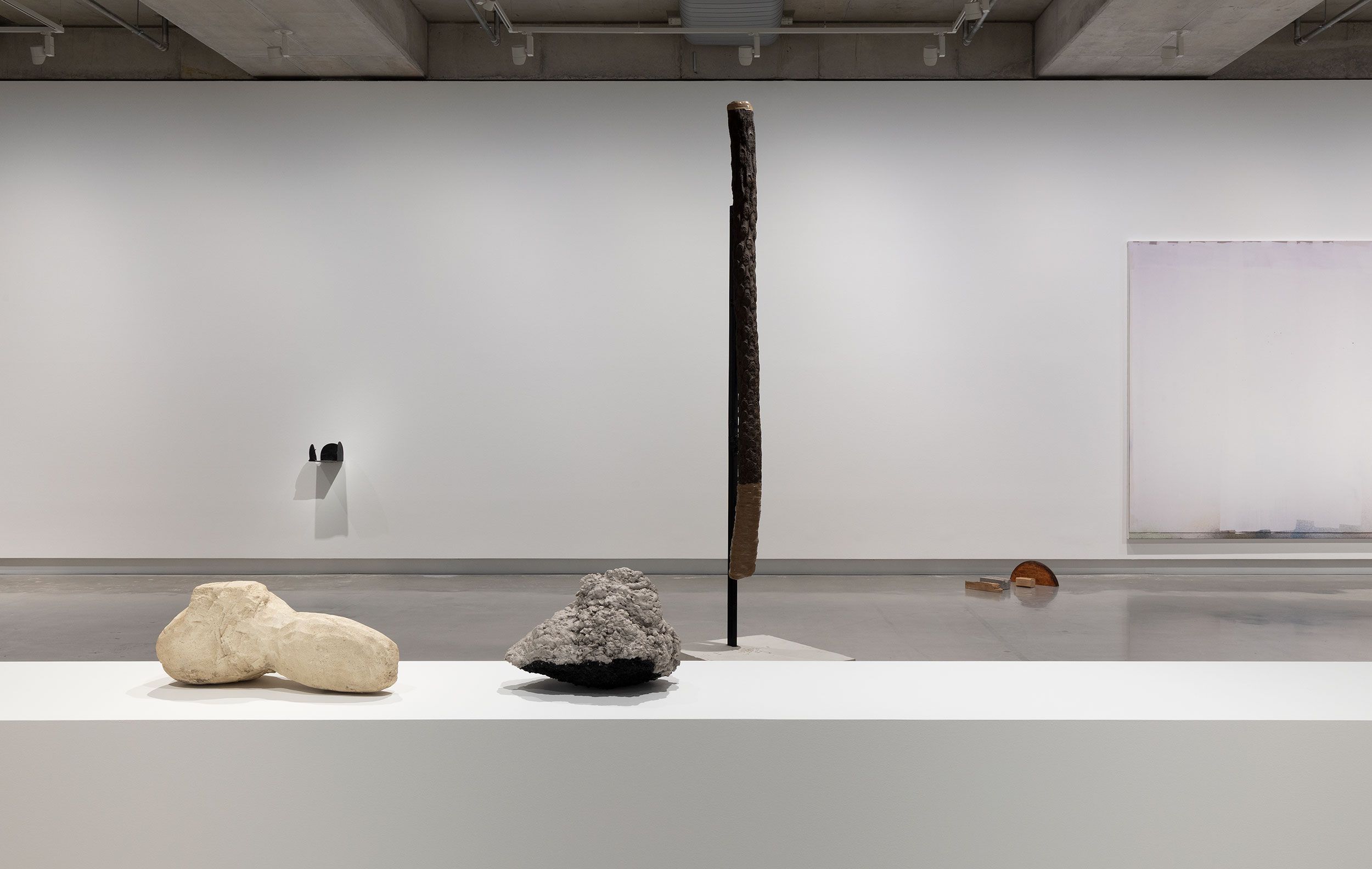 A contemporary art gallery with minimal sculptures: two organic-shaped stones on a white pedestal, a tall vertical sculpture in the background, a wall-mounted piece, and an abstract artwork on the far right wall.