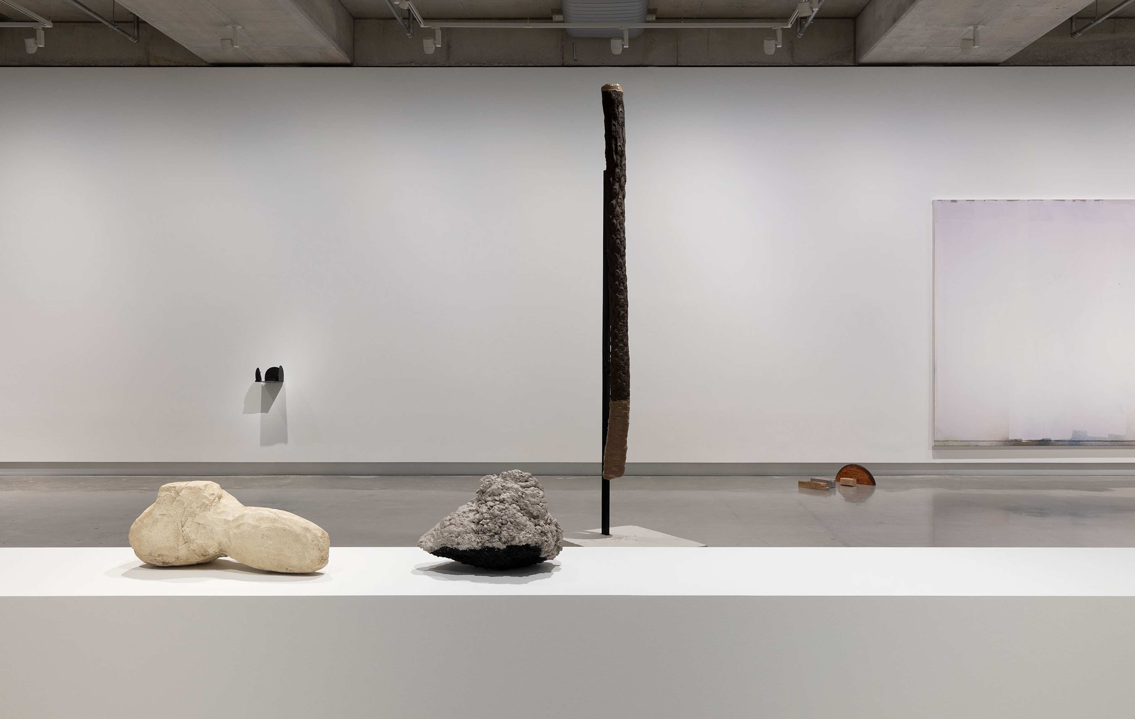 A contemporary art gallery with minimal sculptures: two organic-shaped stones on a white pedestal, a tall vertical sculpture in the background, a wall-mounted piece, and an abstract artwork on the far right wall.