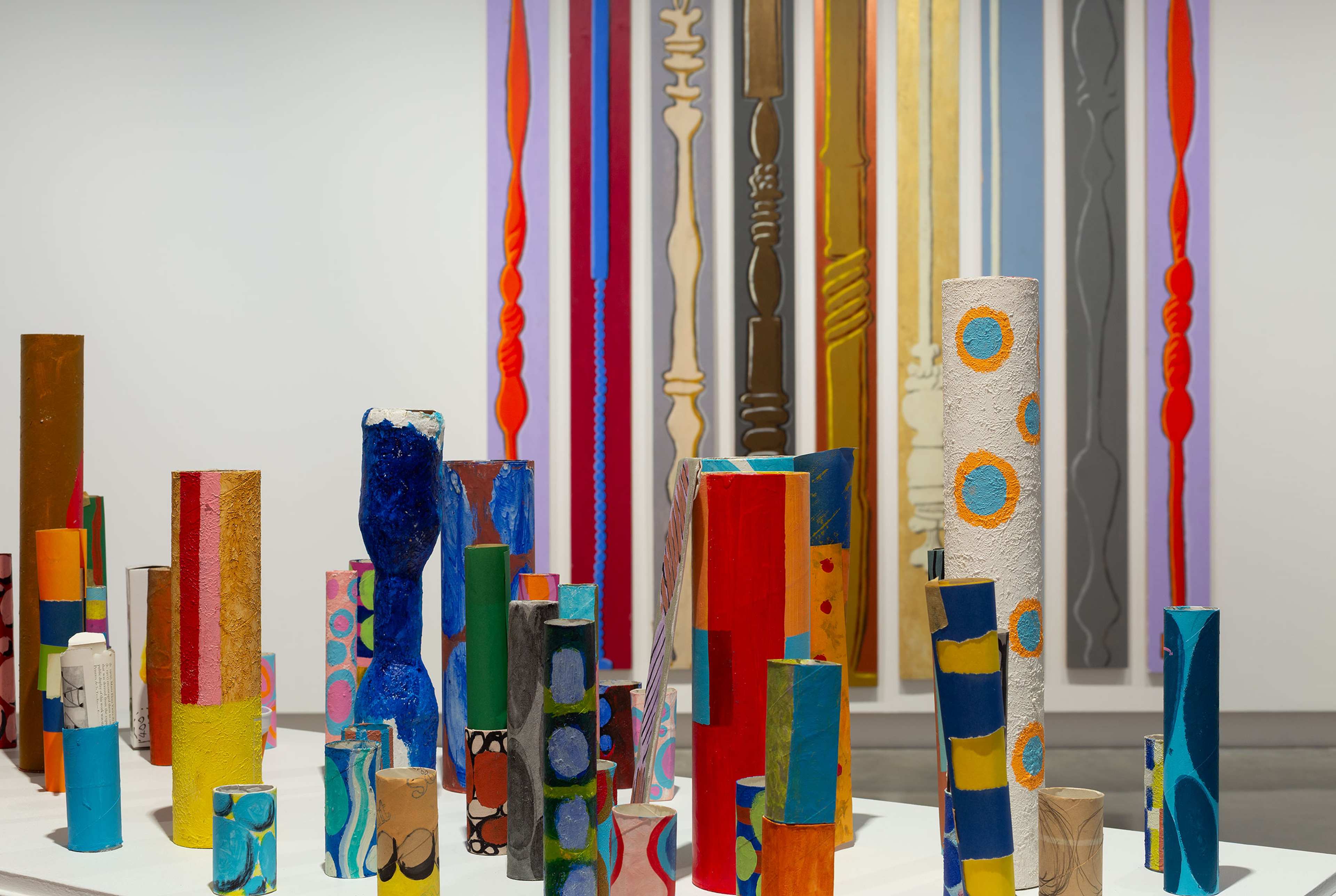 Colourful painted wooden columns of various heights are displayed on white platforms, with tall, vertically striped and patterned wall art in the background, creating a vibrant, abstract art installation in a gallery space.