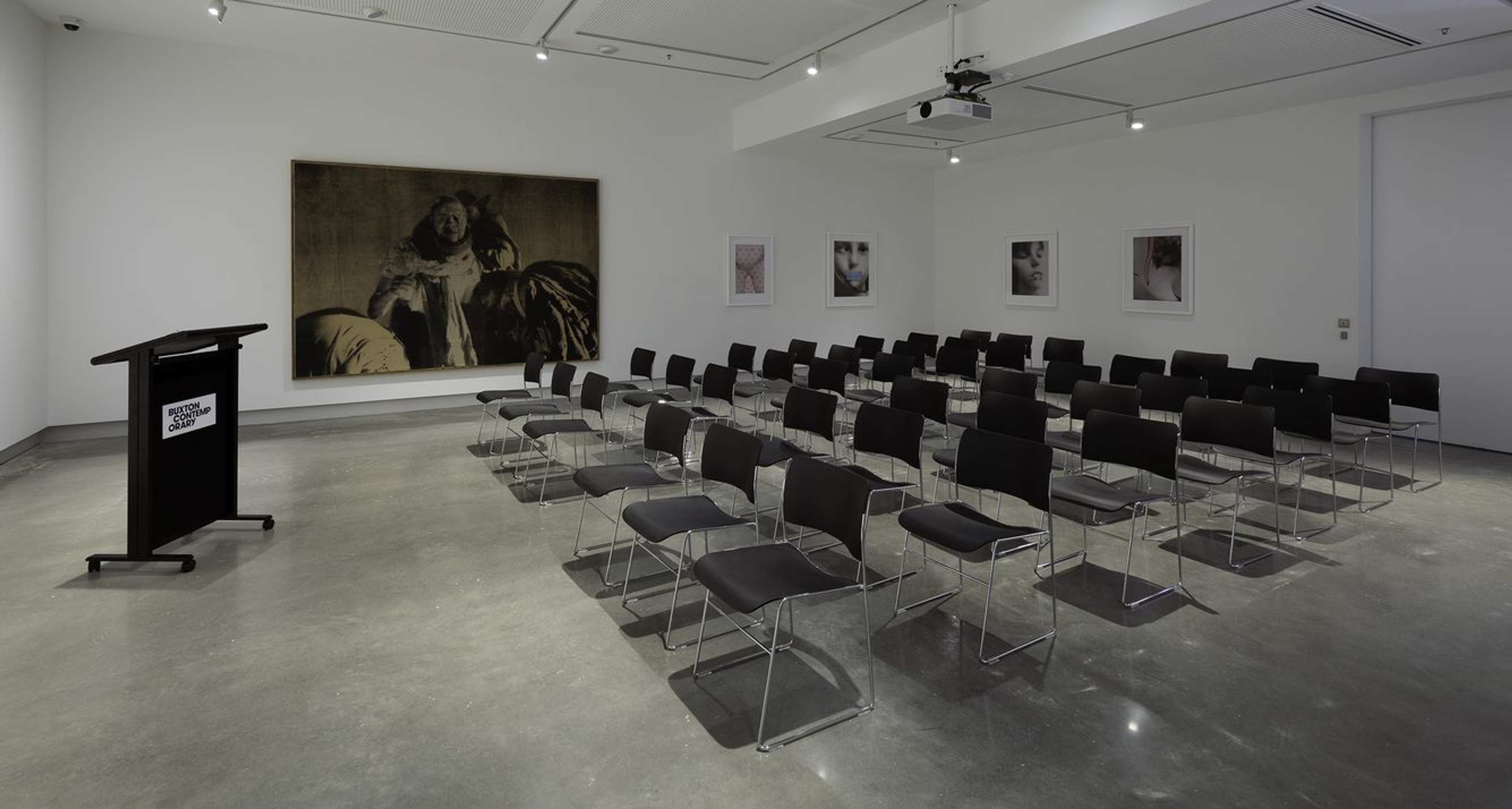 A modern conference room with rows of empty black chairs facing a podium on the left. Artworks hang on the white walls, and a projector is mounted on the ceiling. The floor is polished concrete.