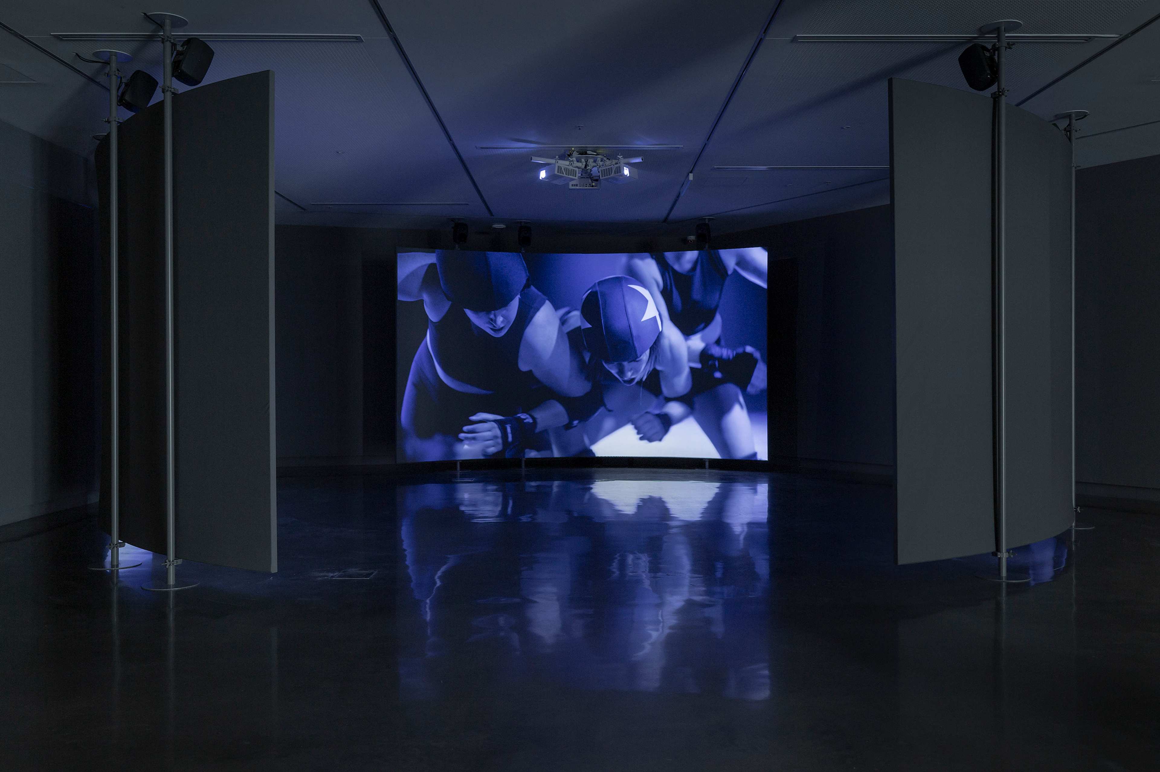A dark gallery features a large screen displaying three athletes in helmets and uniforms, appearing to wrestle or play a contact sport. Panels curve towards the screen, and the room is dimly lit with blue tones reflected on the shiny floor.
