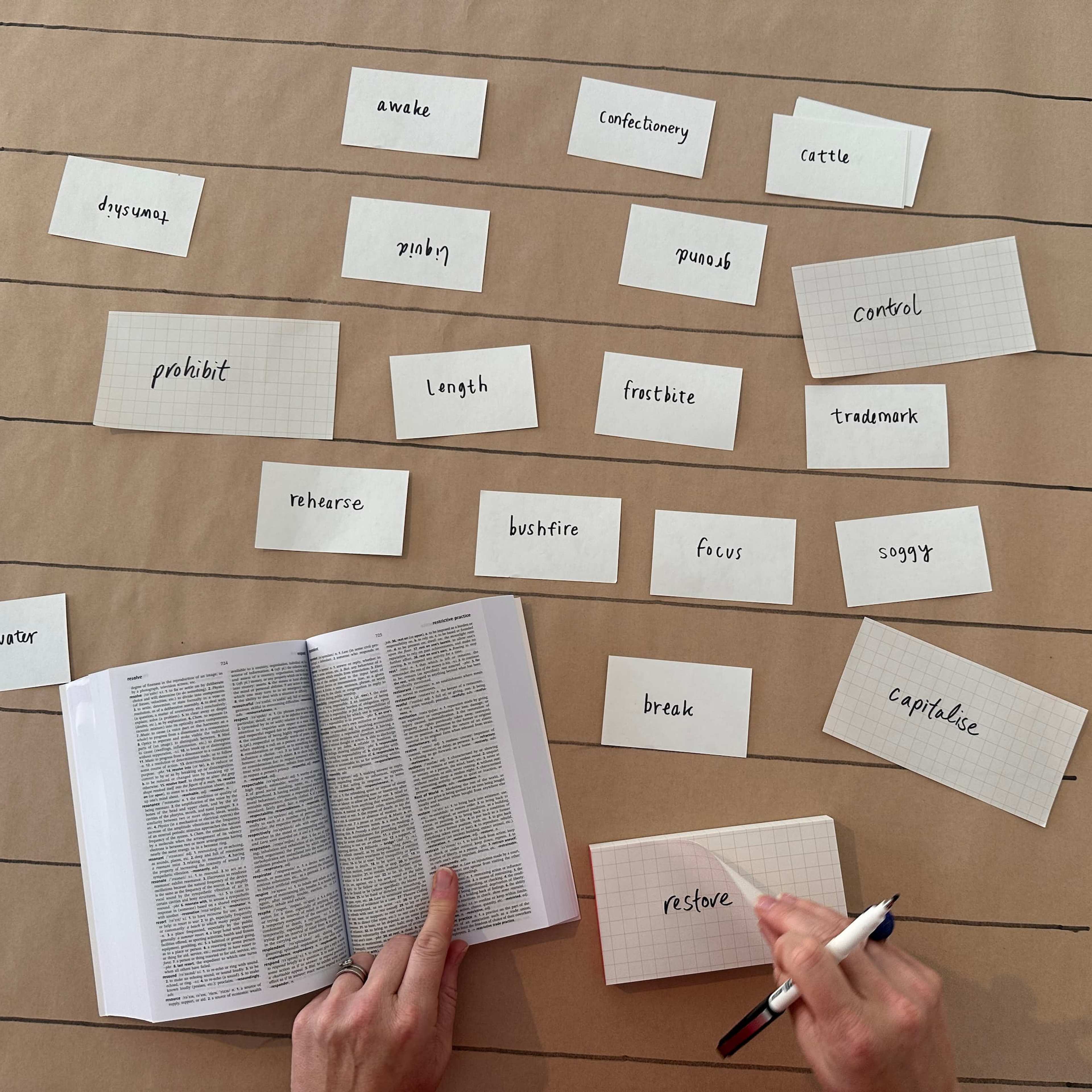 A person writes on an index card while looking at an open book. Several note cards with words like control, prohibit, and focus are arranged in rows on a brown paper-covered table.