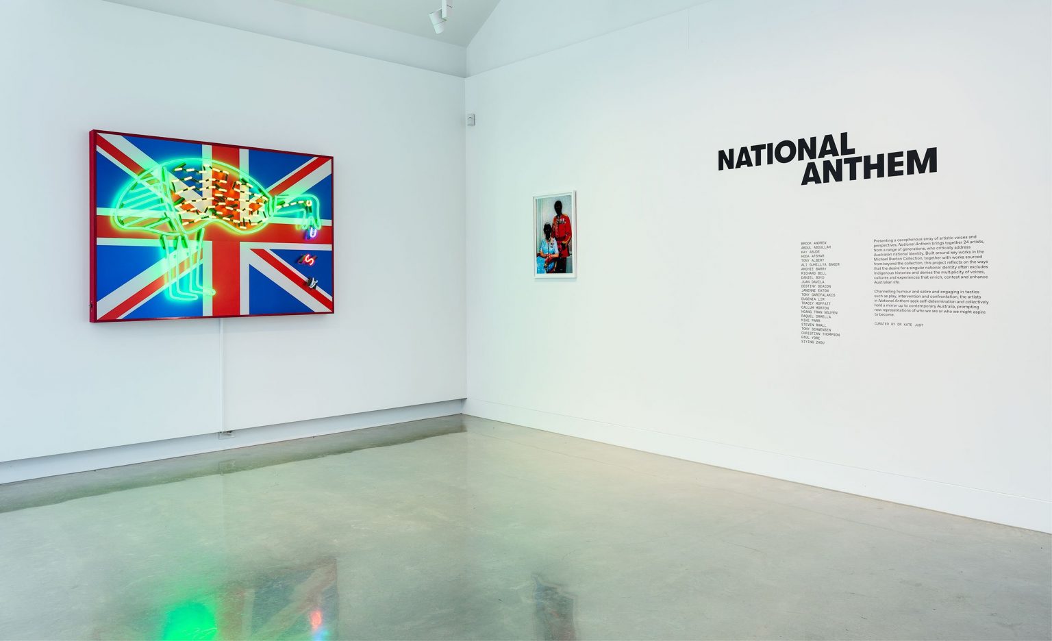 A modern art gallery with a neon artwork of a bird on a Union Jack flag, and the title NATIONAL ANTHEM on the wall beside text and a small photo of a person in red clothing. The gallery floor is shiny and light gray.