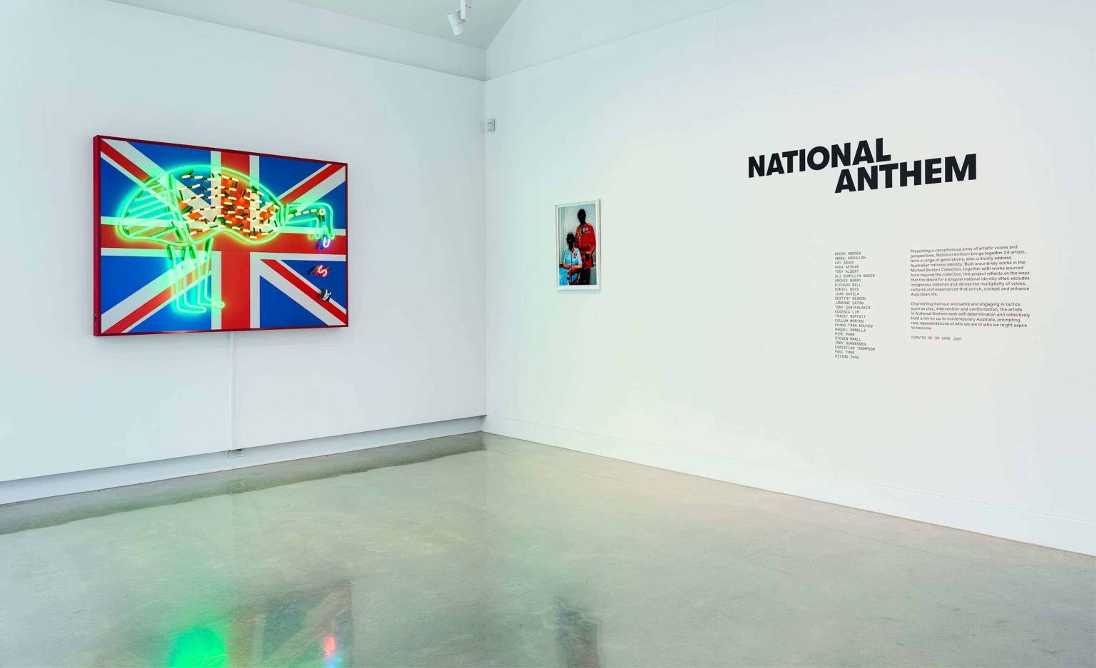 A modern art gallery with a neon artwork of a bird on a Union Jack flag, and the title NATIONAL ANTHEM on the wall beside text and a small photo of a person in red clothing. The gallery floor is shiny and light gray.