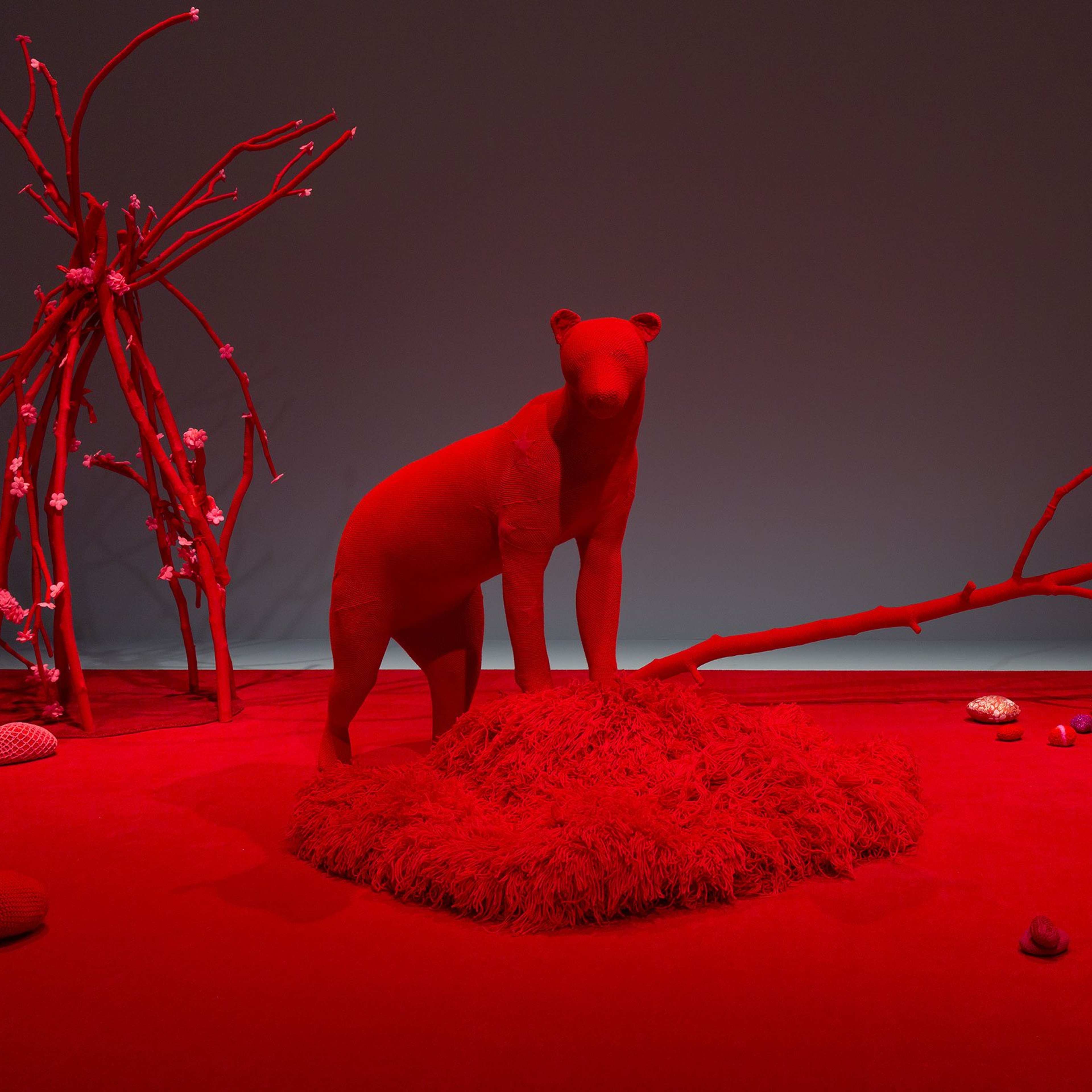 A surreal red-lit scene features a bear-like statue on a shaggy mound, bare branches, scattered stones and shells, and abstract shapes on a red floor and background, creating an otherworldly atmosphere.