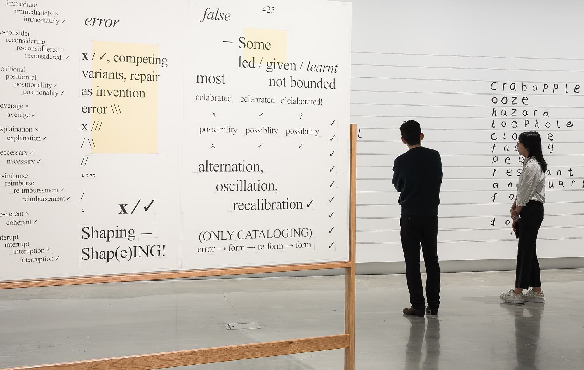 Two people observe large white panels covered with black text, diagrams, and highlighted phrases in an art gallery with polished concrete floors and modern white walls.