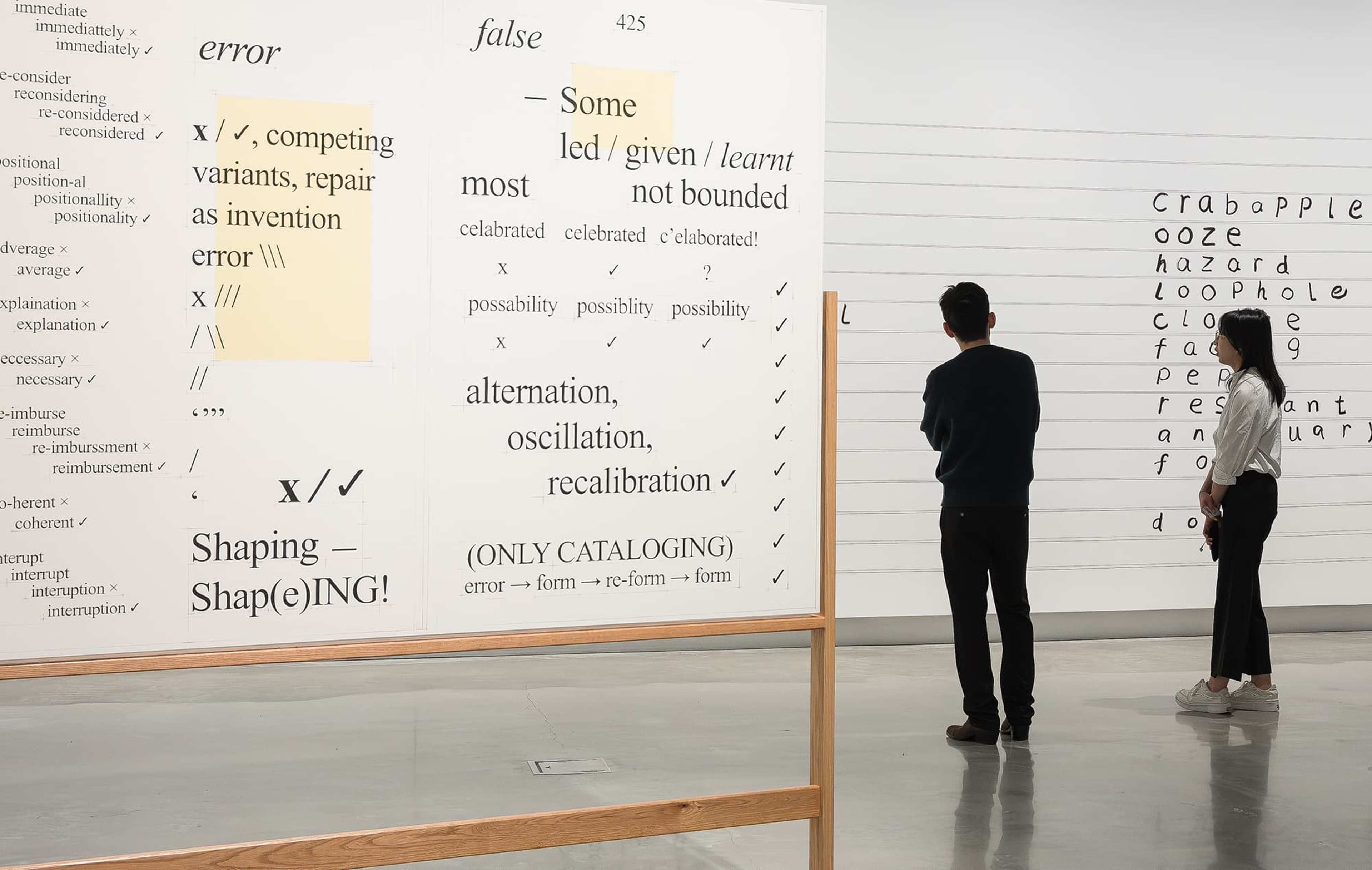 Two people observe large white panels covered with black text, diagrams, and highlighted phrases in an art gallery with polished concrete floors and modern white walls.