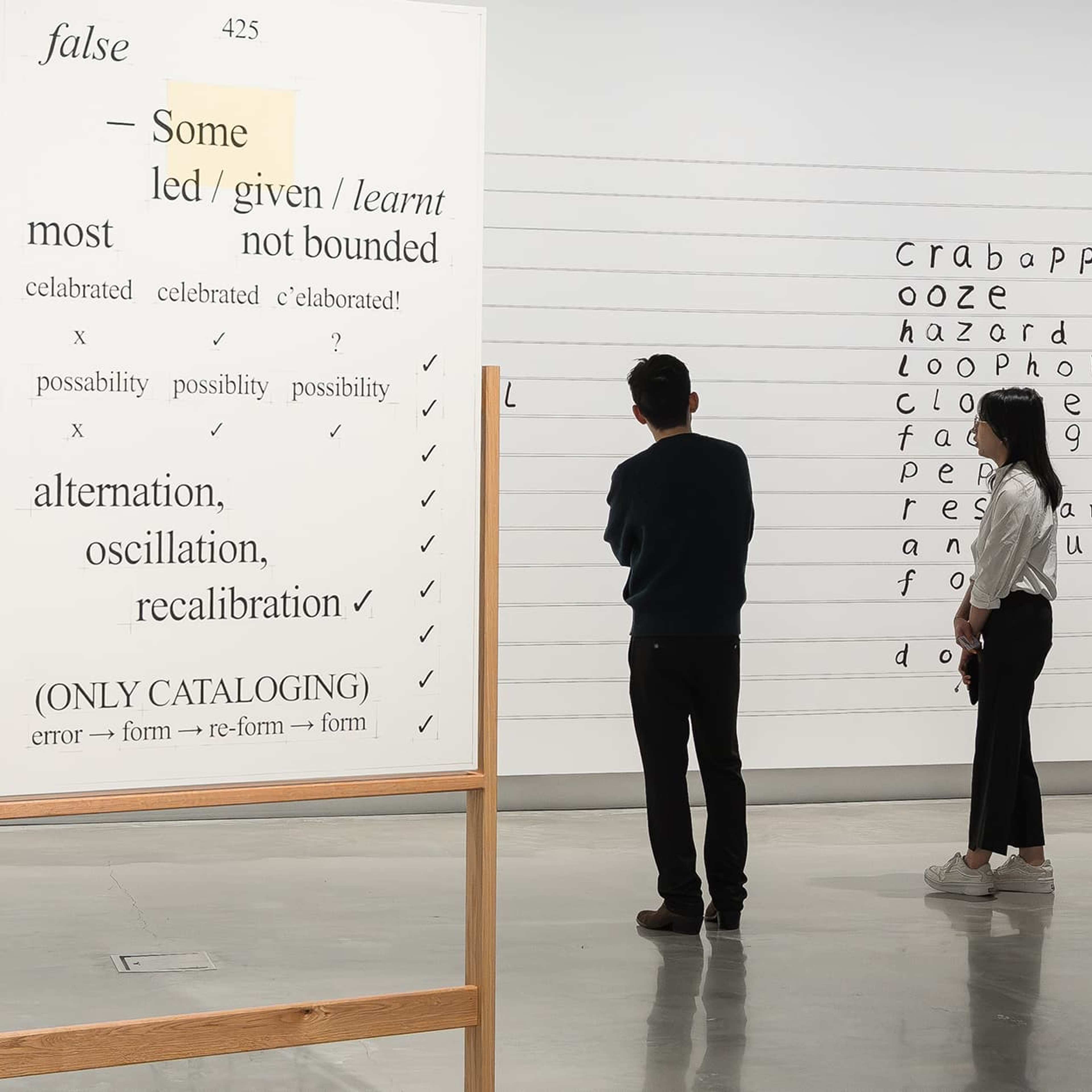 Two people observe large white panels covered with black text, diagrams, and highlighted phrases in an art gallery with polished concrete floors and modern white walls.