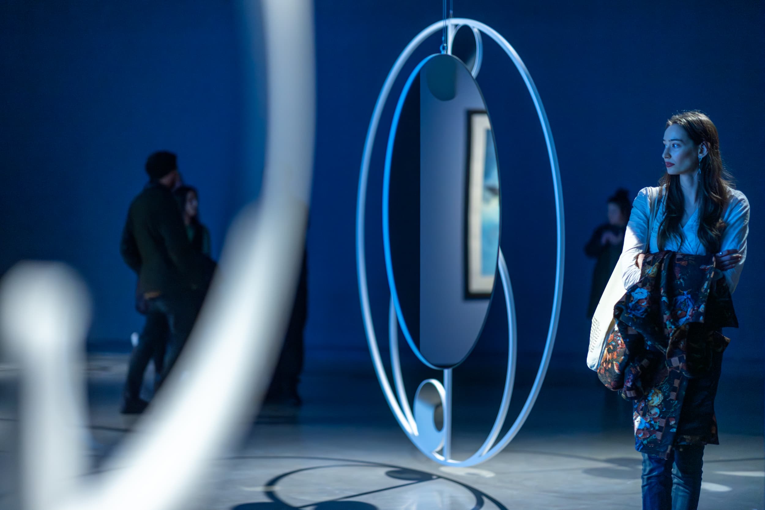 A woman stands in a gallery looking thoughtfully at modern circular sculptures; people are seen blurred in the background under blue lighting.