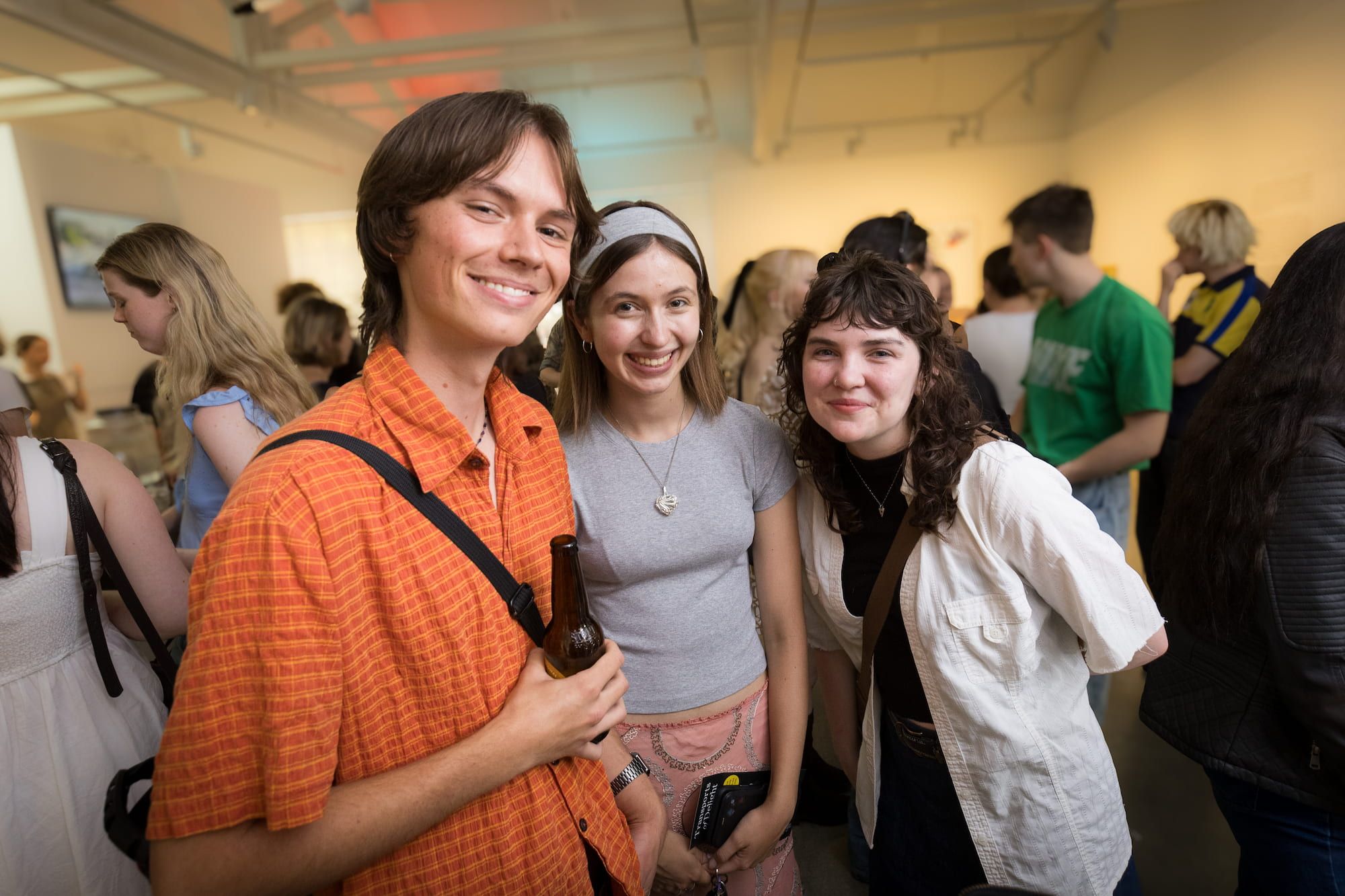 Three young adults smile at the camera at a social gathering in a brightly lit room, surrounded by other people mingling in the background. One person holds a drink.