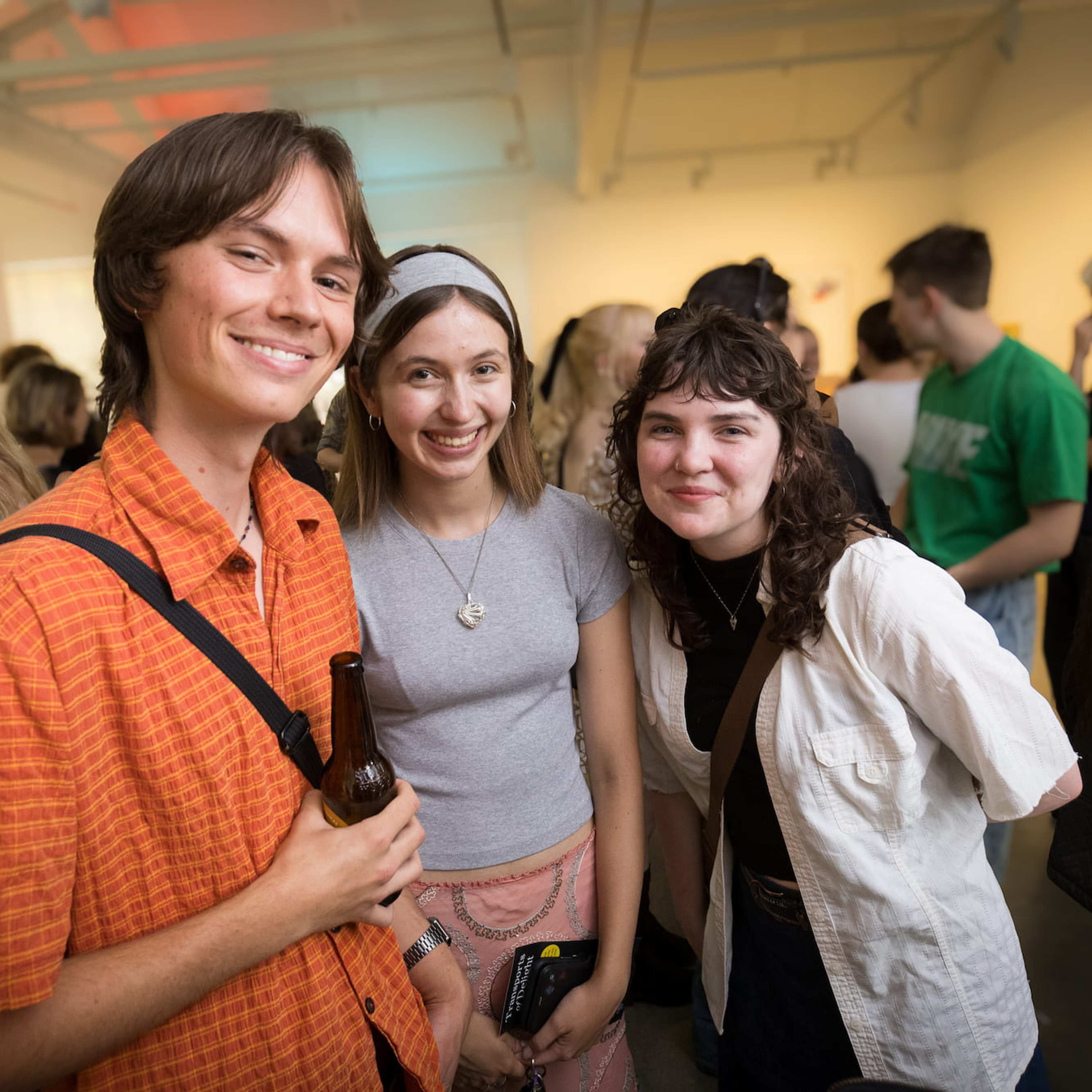 Three young adults smile at the camera at a social gathering in a brightly lit room, surrounded by other people mingling in the background. One person holds a drink.