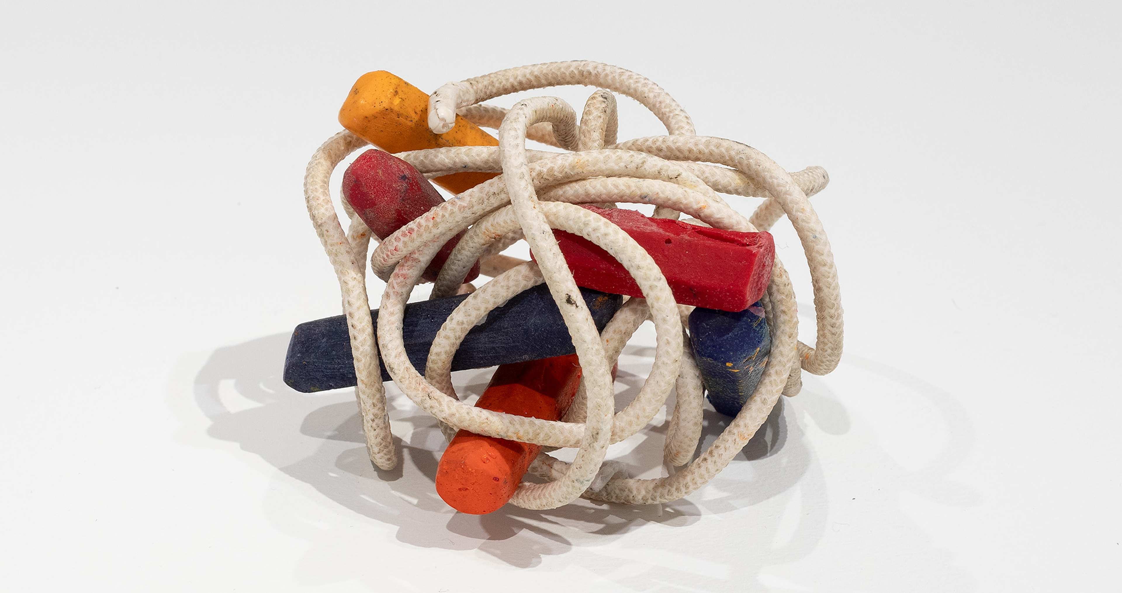 A tangle of white rope wraps around several colorful wooden sticks—red, orange, yellow, and blue—arranged in a loose, intertwined cluster on a white background.
