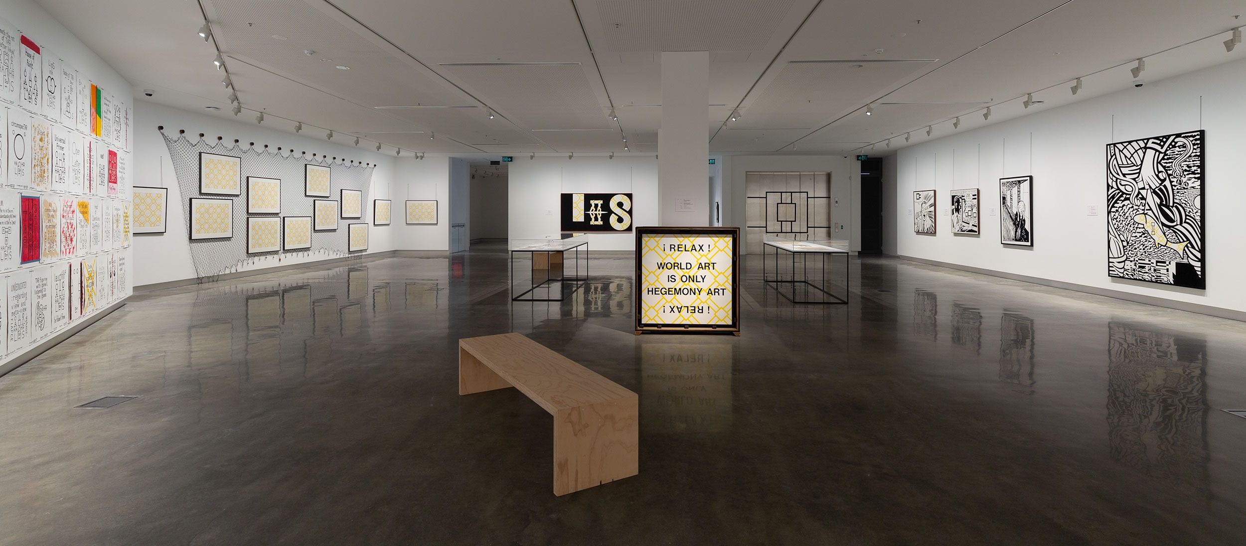 A contemporary art gallery with white walls displays graphic black-and-white artworks and text-based pieces. A wooden bench sits in the center, and a light-up sign reads, I REALLY WISH ART IS ONLY DESIGN ART. The floor is polished concrete.