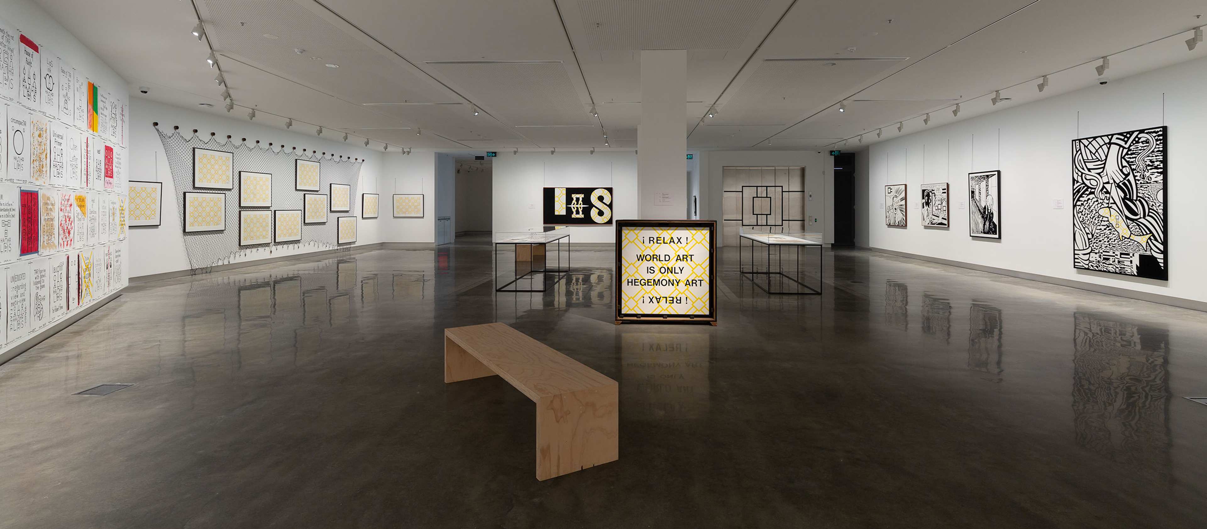 A contemporary art gallery with white walls displays graphic black-and-white artworks and text-based pieces. A wooden bench sits in the center, and a light-up sign reads, I REALLY WISH ART IS ONLY DESIGN ART. The floor is polished concrete.