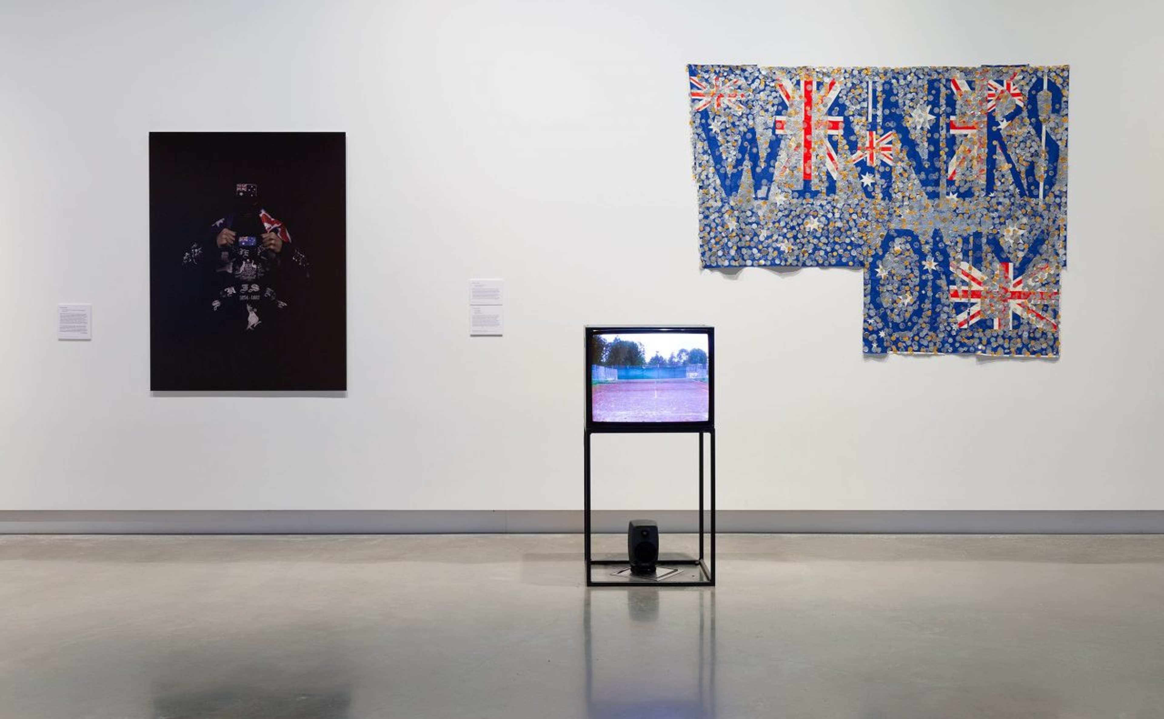A gallery wall displays abstract textile art with Union Jack patterns, a dark framed artwork, and a TV on a stand playing outdoor footage, all set on a polished concrete floor.