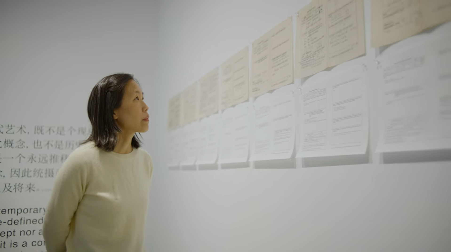 Carol Yinghua Lu looking up at printed paper works hanging on a wall.