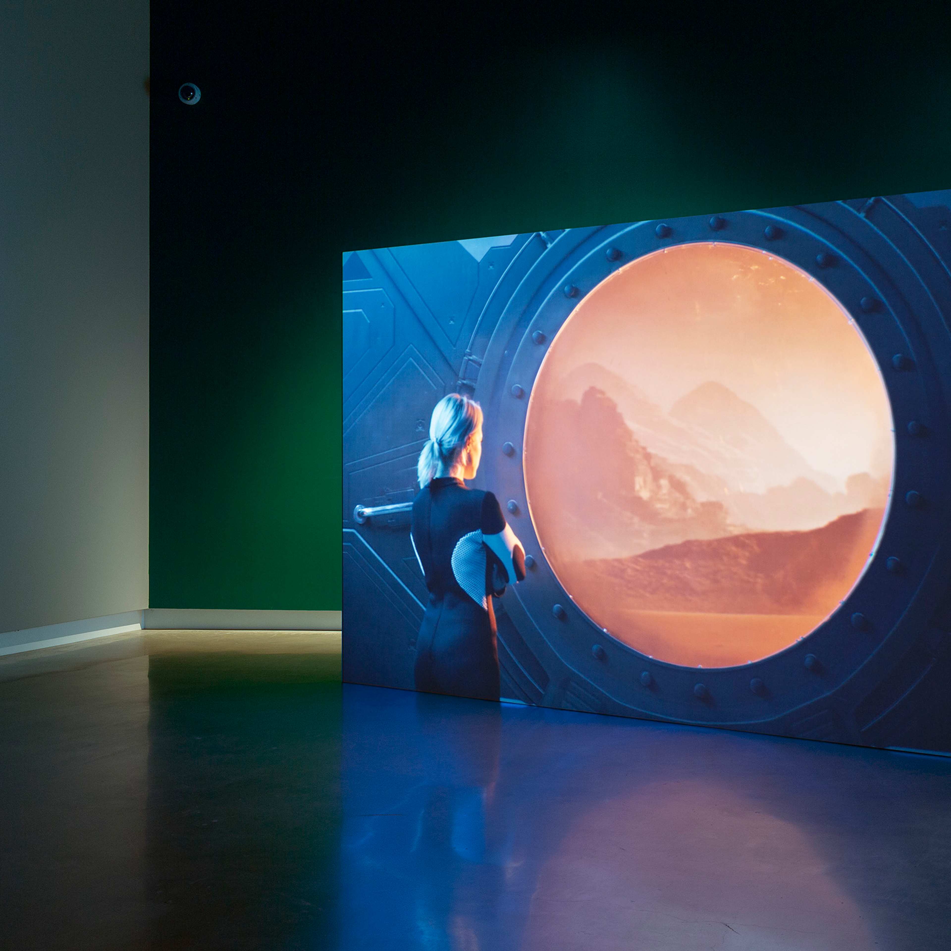 A digital artwork shows a woman with blue hair looking out a large circular window at an orange-hued alien landscape with mountains, displayed against a dark green wall in a gallery space.